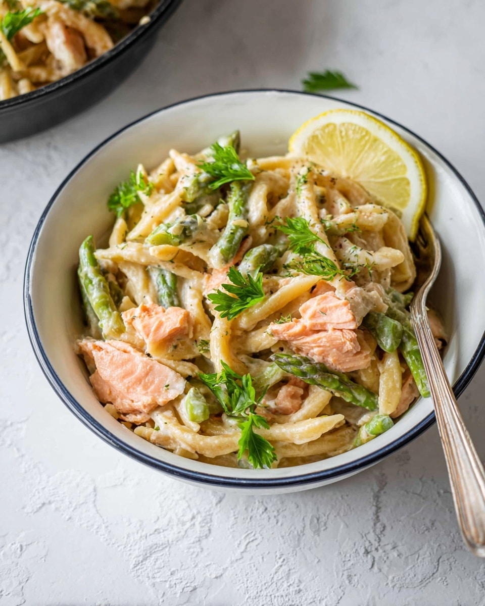 A bowl with creamy pasta layered at the bottom in light beige color, mixed with pieces of cooked salmon in pink-orange shade scattered on top, with bright green asparagus pieces placed throughout the dish. Fresh green parsley leaves are sprinkled on the pasta, and a quarter slice of lemon with yellow skin and pale inner flesh is placed on the edge. A silver fork rests inside the bowl on the right side. The bowl is white with a thin blue rim, sitting on a white marbled surface. Photo taken with an iphone --ar 4:5 --v 7