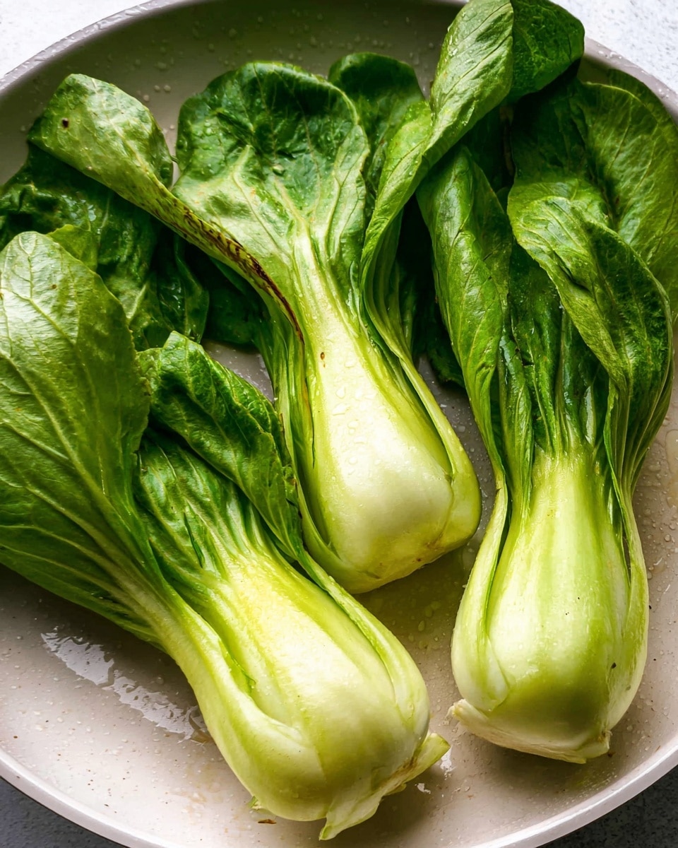 Four pieces of fresh bok choy with bright green, slightly wrinkled leaves and pale, smooth bulbs are laid out in a round white pan with slight water droplets visible. The bok choy leaves show some darkened edges, and the bulbs have a few light brown spots and a firm texture. The white marbled surface of the background enhances the freshness of the bok choy. photo taken with an iphone --ar 4:5 --v 7