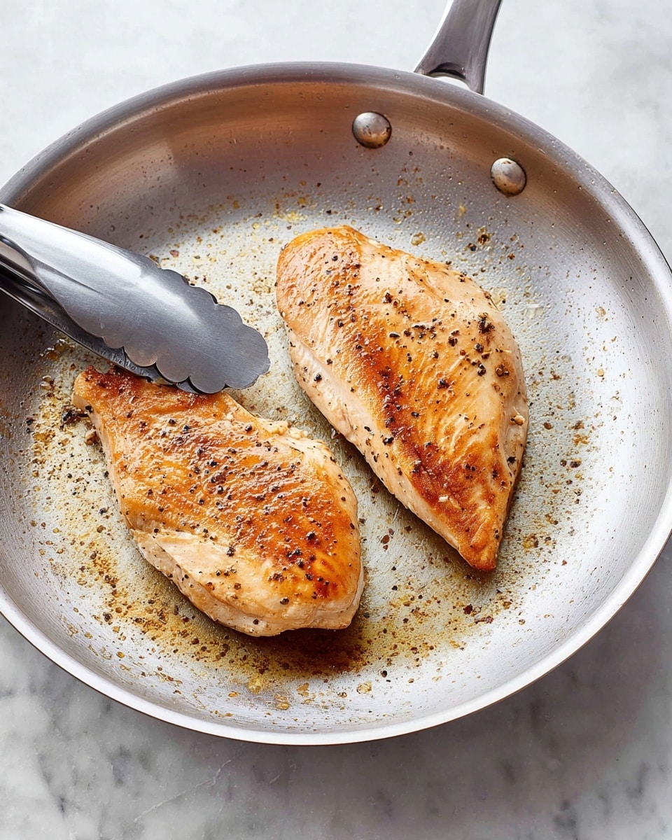 Two cooked chicken fillets with a light brown and slightly crispy surface lie flat on a silver metal frying pan. The fillets have a golden-brown sear with small black pepper specks and some light char marks. The pan shows bits of browned residue around the fillets from cooking, giving it a textured look. A silver tong with black grip holds the fillet on the top left side. The pan rests on a white marbled surface. photo taken with an iphone --ar 4:5 --v 7