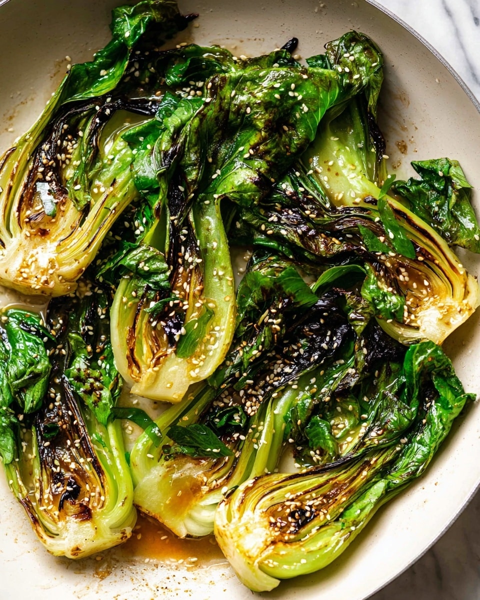 A white pan holds four pieces of cooked bok choy, each sliced in half lengthwise. The bok choy is bright green with parts that are darkened and charred from cooking, showing a mix of light green and white at the thick base and vibrant dark green leaves curling at the top. Small sesame seeds are sprinkled evenly over the bok choy, adding texture and contrast. The pan has a slightly oily shine with some browned bits from cooking, and the background is a white marbled texture. photo taken with an iphone --ar 4:5 --v 7