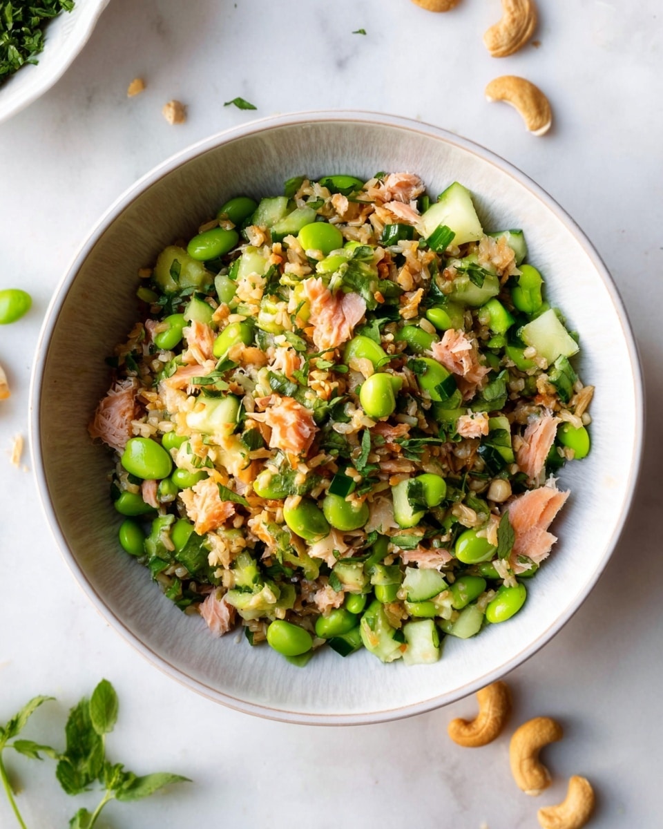 A bowl with one main layer of mixed ingredients including bright green edamame beans, light and dark green cucumber pieces, small chunks of pink cooked fish, and light brown cooked rice, all evenly combined. The mixture has small bits of green herbs sprinkled throughout, adding texture and color contrast. Around the bowl, on a white marbled surface, are some scattered cashews and fresh green herb leaves visible at the edge. The bowl itself is white with a subtle light pattern on the inside rim. Photo taken with an iphone --ar 4:5 --v 7