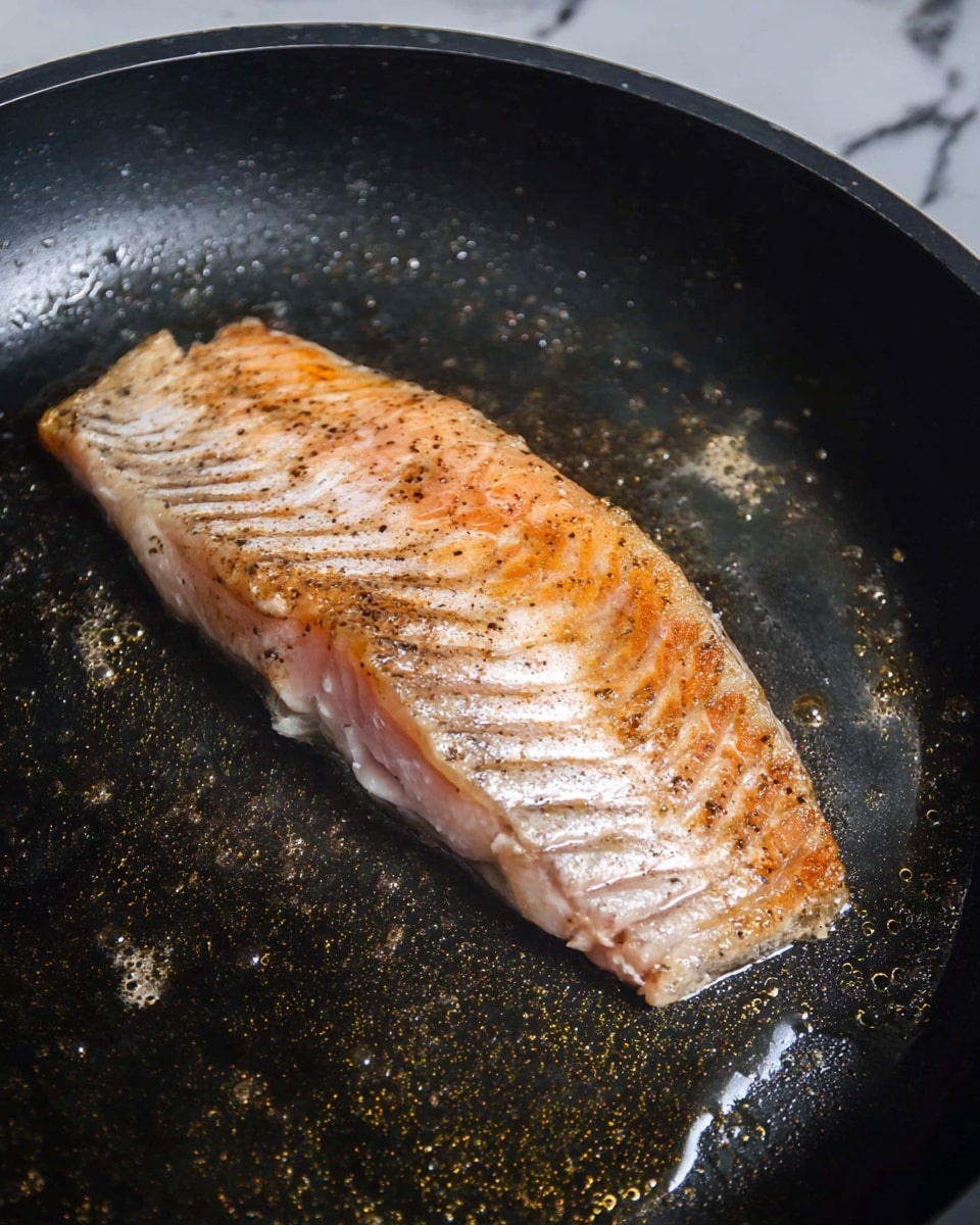 A single piece of fish fillet is cooking in a black frying pan. The fillet has two layers: the top layer is golden brown with a crispy texture and some black pepper sprinkled over it, while the bottom layer shows light pink moist flesh. The pan has small amounts of oil and some water droplets around the fillet. The background shows a white marbled texture. photo taken with an iphone --ar 4:5 --v 7