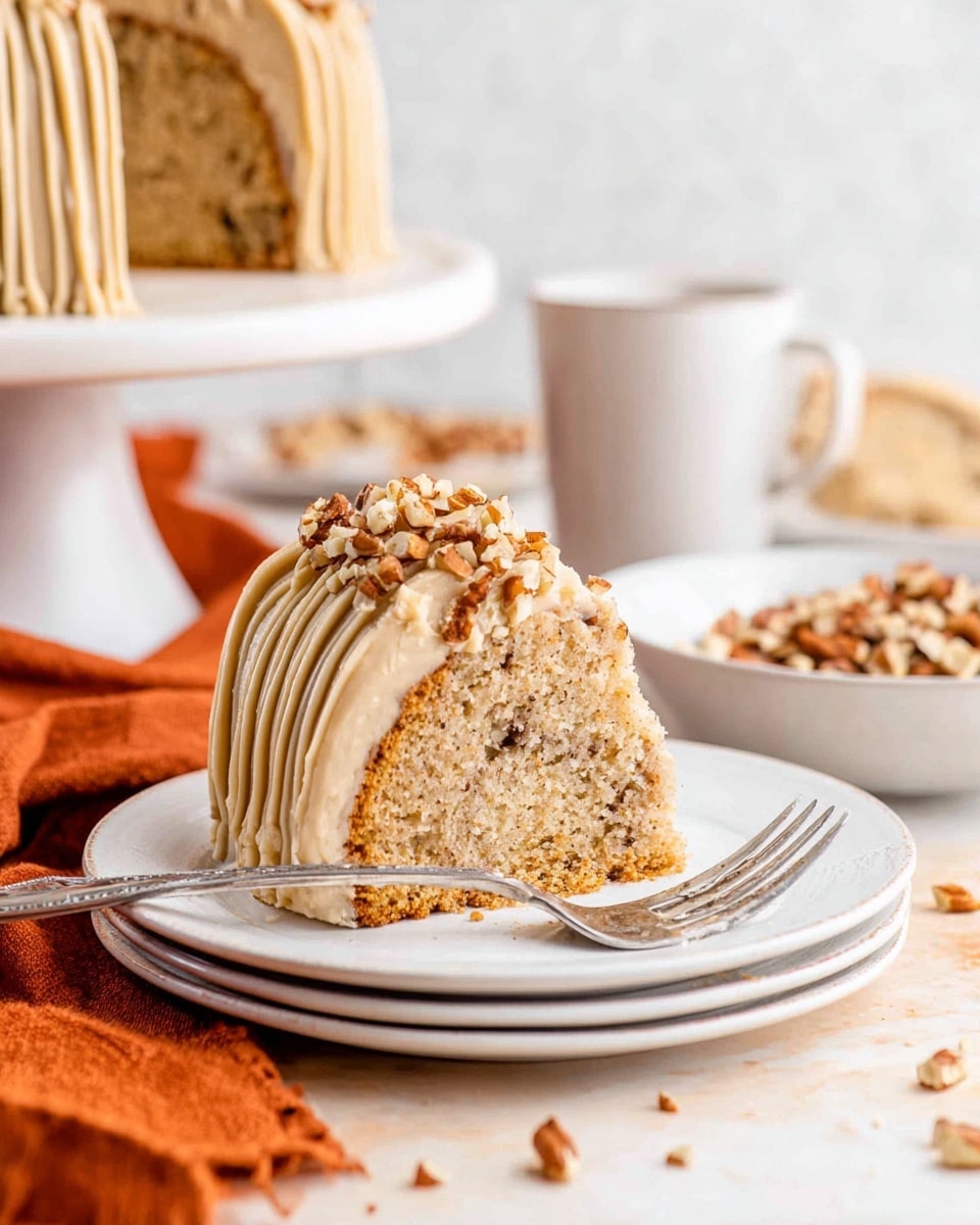 A single slice of light brown cake with small dark nut pieces inside sits on a white plate. The cake has one thick layer, topped with creamy light tan frosting that is piped in vertical lines around the side and on top. Chopped nuts are sprinkled over the top of the frosting. A silver fork rests beside the slice on the plate. In the background, there is an orange cloth on a white marbled surface, a white bowl filled with more nuts, and a white mug and plate with more cake slices, all softly out of focus. photo taken with an iphone --ar 4:5 --v 7