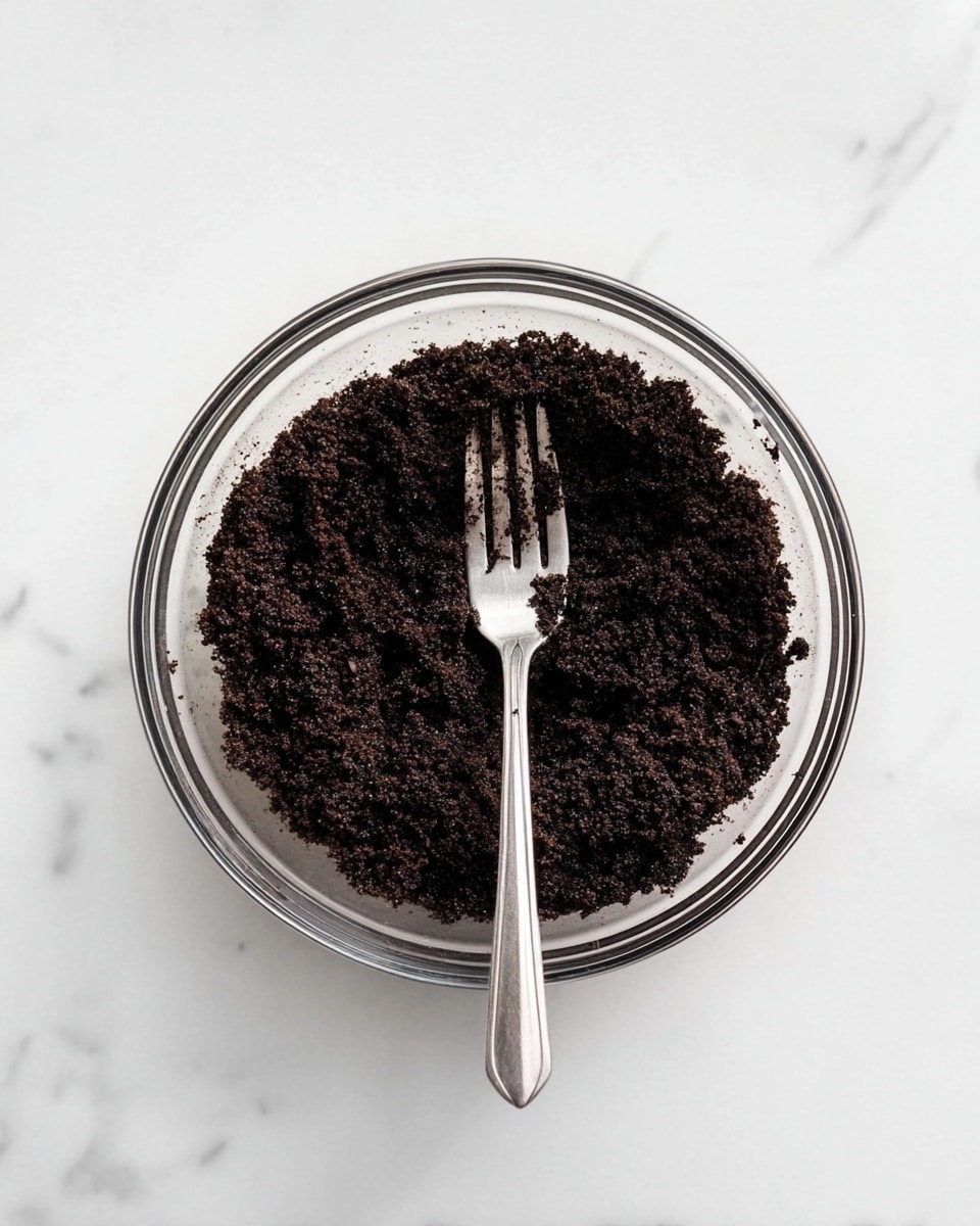 A clear glass bowl filled with a layer of dark brown crumbly mixture, likely crushed cookies or cake crumbs, resting on a white marbled surface. In the center of the bowl, a silver fork is placed vertically, with some crumbs sticking to its tines, adding texture to the image. The scene is simple and focused on the single bowl and its contents, captured from a top-down view. photo taken with an iphone --ar 4:5 --v 7
