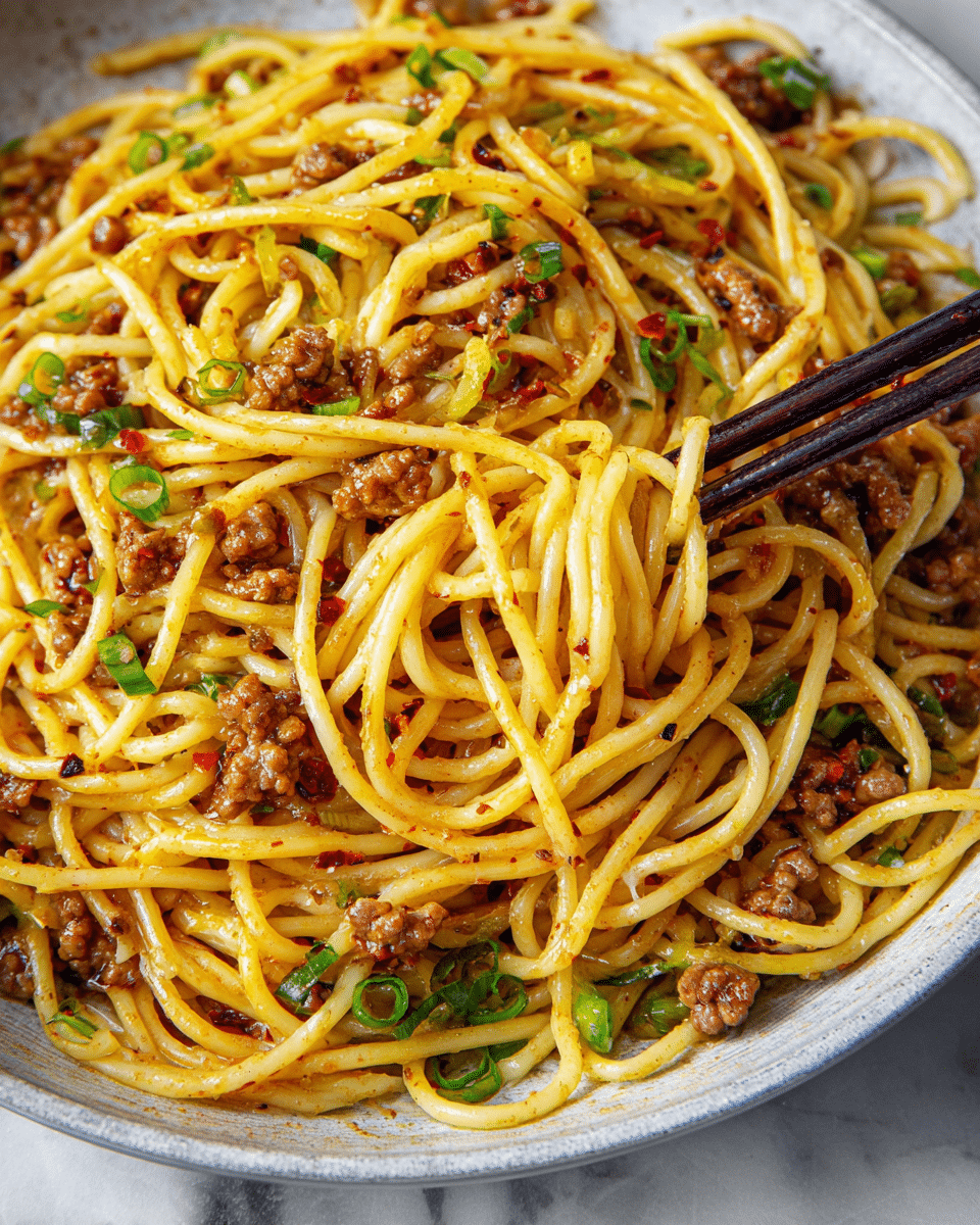 A close-up view of a plate filled with yellow spaghetti noodles mixed with small brown pieces of cooked meat and scattered green onion slices. The noodles are shiny and coated with a light red chili oil sauce, giving a glistening effect, and some red chili flakes are visible throughout. Dark wooden chopsticks are picking up a small bunch of noodles from the right side. The plate is white and sits on a white marbled surface. Photo taken with an iphone --ar 4:5 --v 7