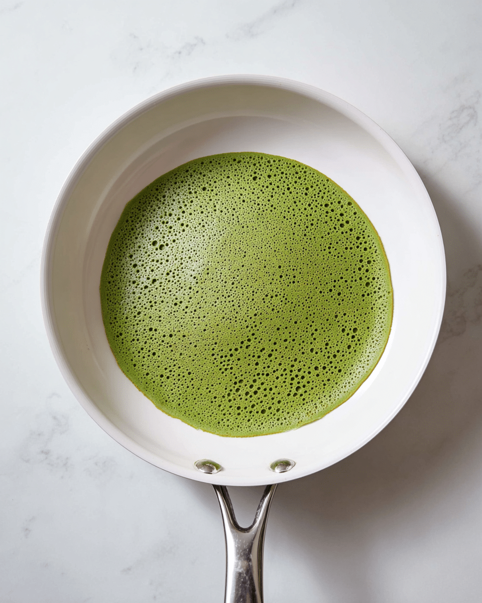 A single round green pancake with many small holes on its surface is cooking in a white pan. The pancake is flat, smooth but dotted, showing a light, airy texture. The white pan has a metal handle and sits on a white marbled surface. photo taken with an iphone --ar 4:5 --v 7