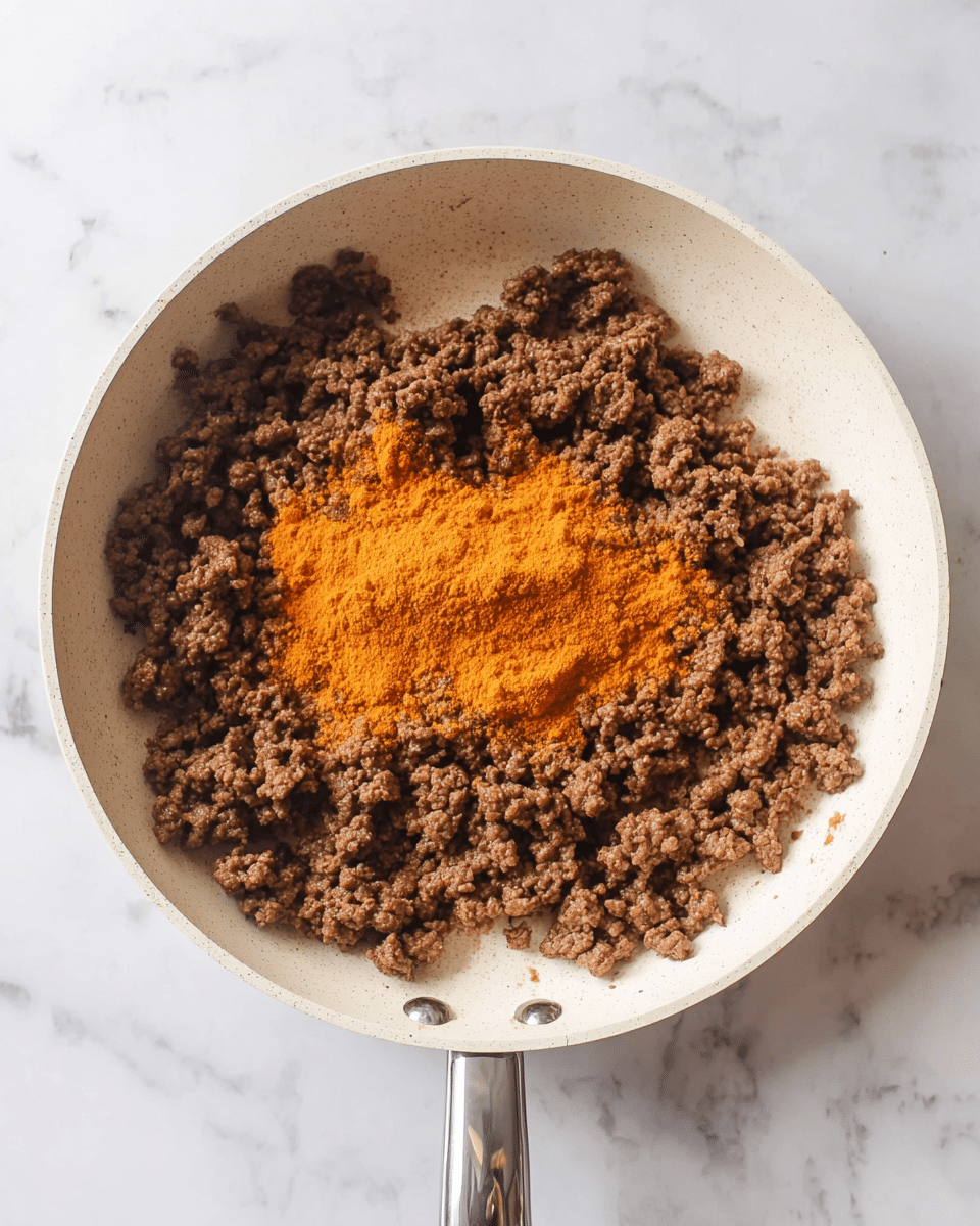 A white frying pan filled with cooked ground beef that is brown and crumbly, covering most of the pan's surface. In the center of the pan, there is a heap of orange-colored powdered spices sitting on top of the beef. The pan rests on a white marbled surface, and the handle extends towards the bottom of the image. photo taken with an iphone --ar 4:5 --v 7