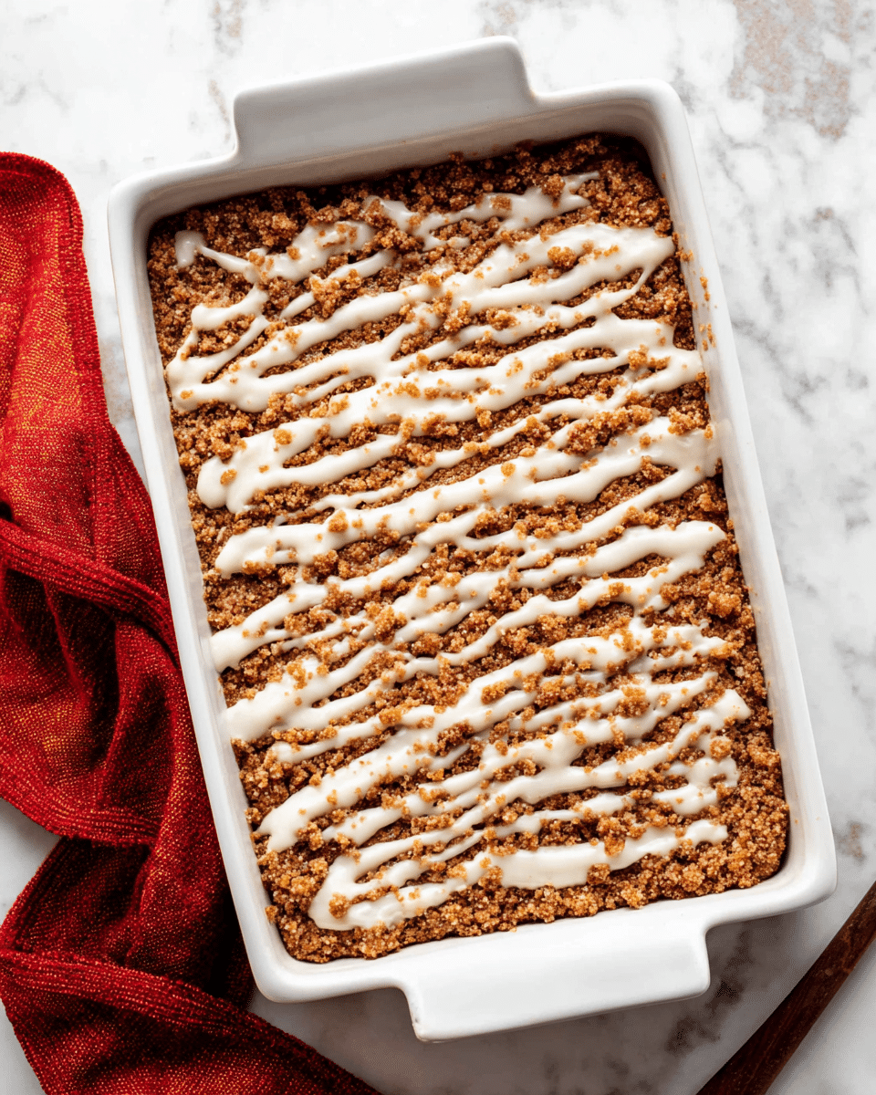 A white rectangular baking dish holds a two-layer dessert. The bottom layer is a dense, crumbly brown base with a rough texture made of small clumps. The top layer is a thin drizzle of white icing spread unevenly across the brown layer, creating a contrast of creamy and crumbly textures. The baking dish is placed on a white marbled surface, and a red cloth is partially visible near the dish. Photo taken with an iphone --ar 4:5 --v 7
