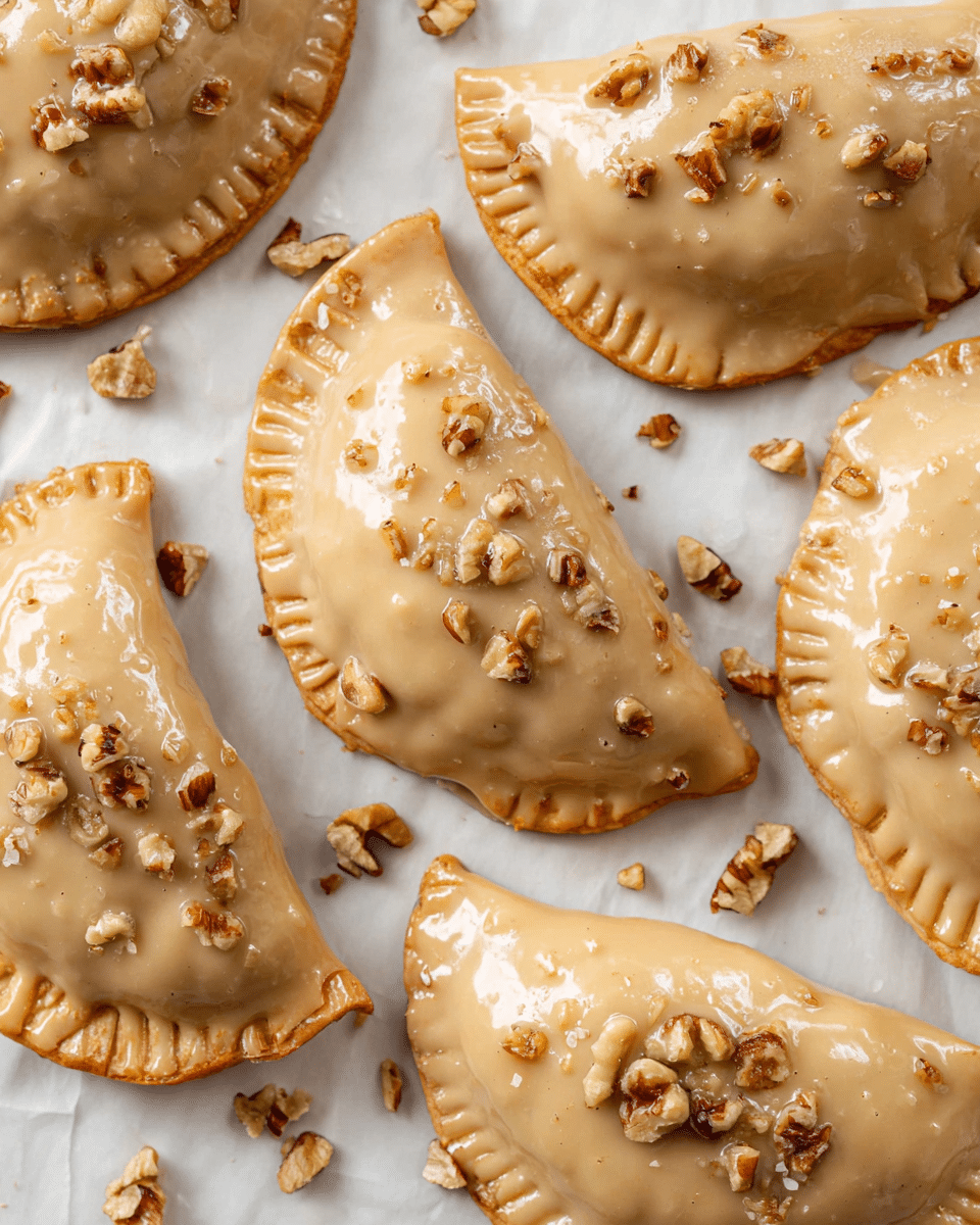 The image shows several crescent-shaped pastries placed on white parchment paper over a white marbled surface. Each pastry is covered in a smooth, light caramel-colored glaze that gives a shiny texture. On top of the glaze, there are small pieces of chopped walnuts scattered evenly across each pastry and a few pieces on the paper around them. The pastries have a slightly crimped edge, showing they were folded and sealed before glazing. The overall color palette is warm beige and brown with textures from the glaze and nut pieces. photo taken with an iphone --ar 4:5 --v 7