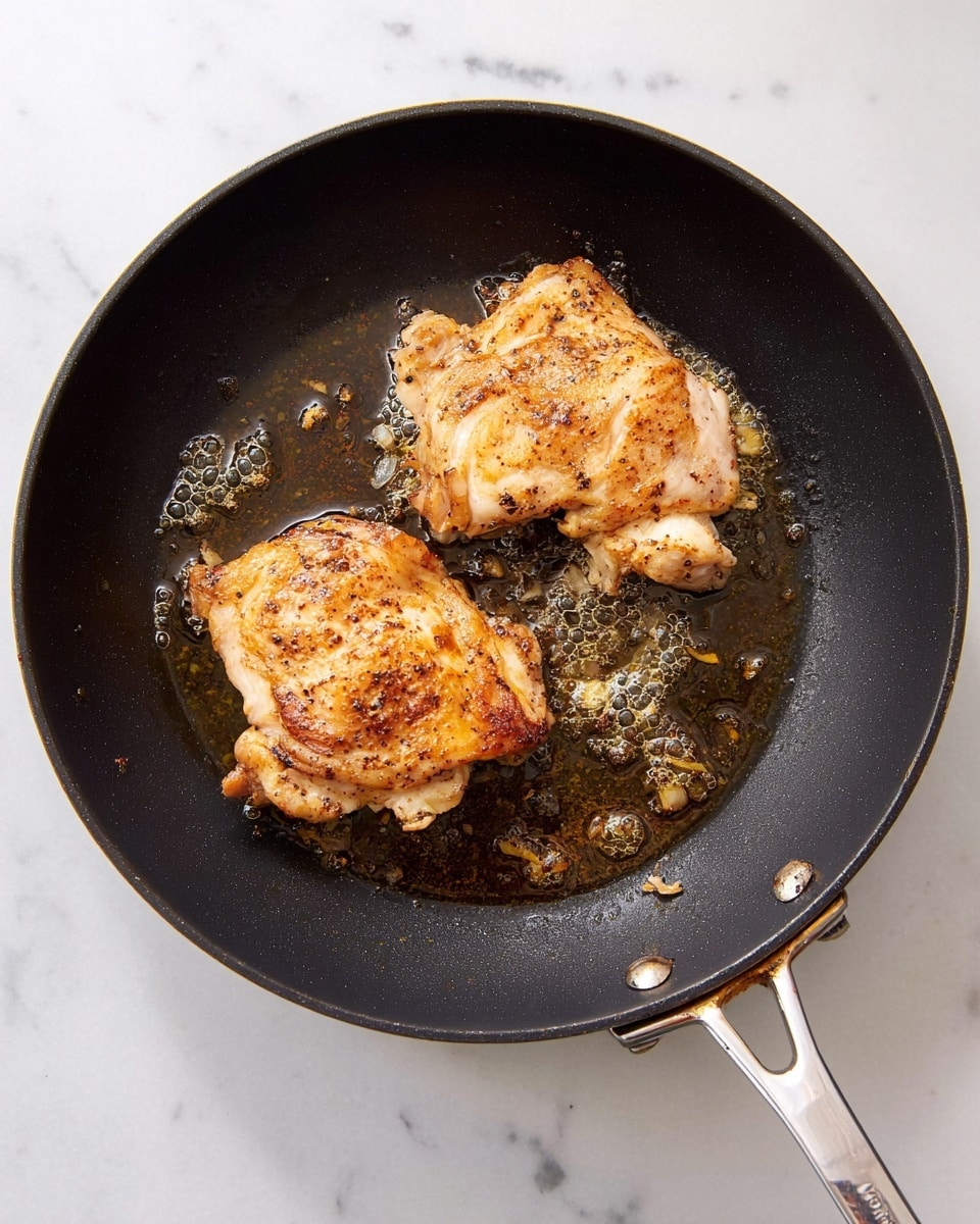 A top view of a black frying pan with two pieces of cooked chicken thighs inside. Each piece has a golden brown and slightly crispy surface with some spots darker due to seasoning, sitting in a thin layer of oil that has bubbles and small bits around. The pan handle is silver and extends out of the frame. The frying pan is placed on a white marbled surface. photo taken with an iphone --ar 4:5 --v 7