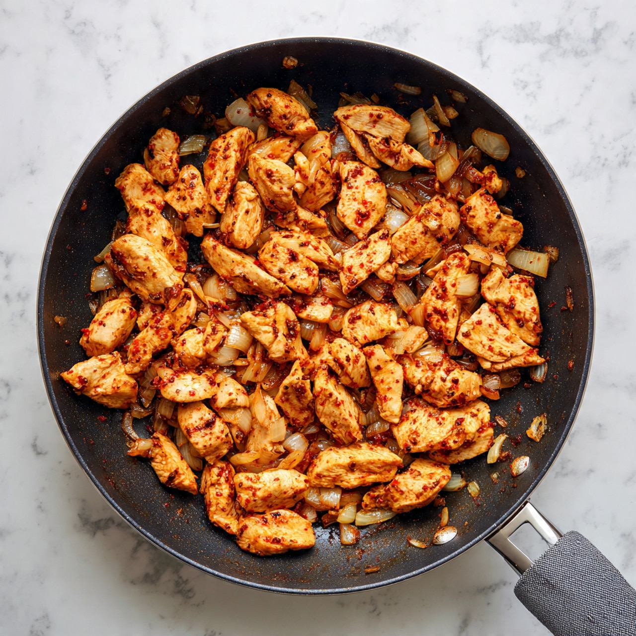 A black frying pan sits on a white marbled surface with cooked chicken strips and small pieces of diced onions inside. The chicken strips are coated with a reddish seasoning and are spread evenly in one layer across the pan, showing some browned edges and a cooked texture. The diced onions are scattered among the chicken pieces, cooked to a translucent light brown. The pan handle is partially visible on the bottom right with a gray cloth nearby. photo taken with an iphone --ar 4:5 --v 7