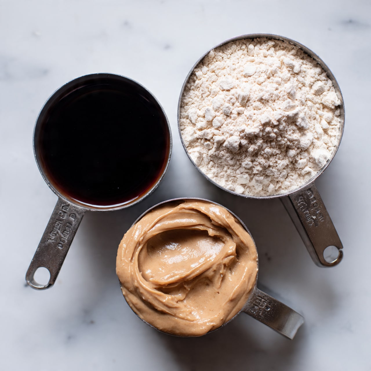 The image shows three measuring cups on a white marbled surface, each filled with a different ingredient. The top right cup contains a pale, powdery flour, with a light and crumbly texture. The bottom right cup is filled with a thick, smooth, light brown creamy substance with a few small lumps, likely peanut butter. The left cup holds a dark, glossy, syrup-like liquid. The cups are arranged loosely triangular with soft natural lighting and the words