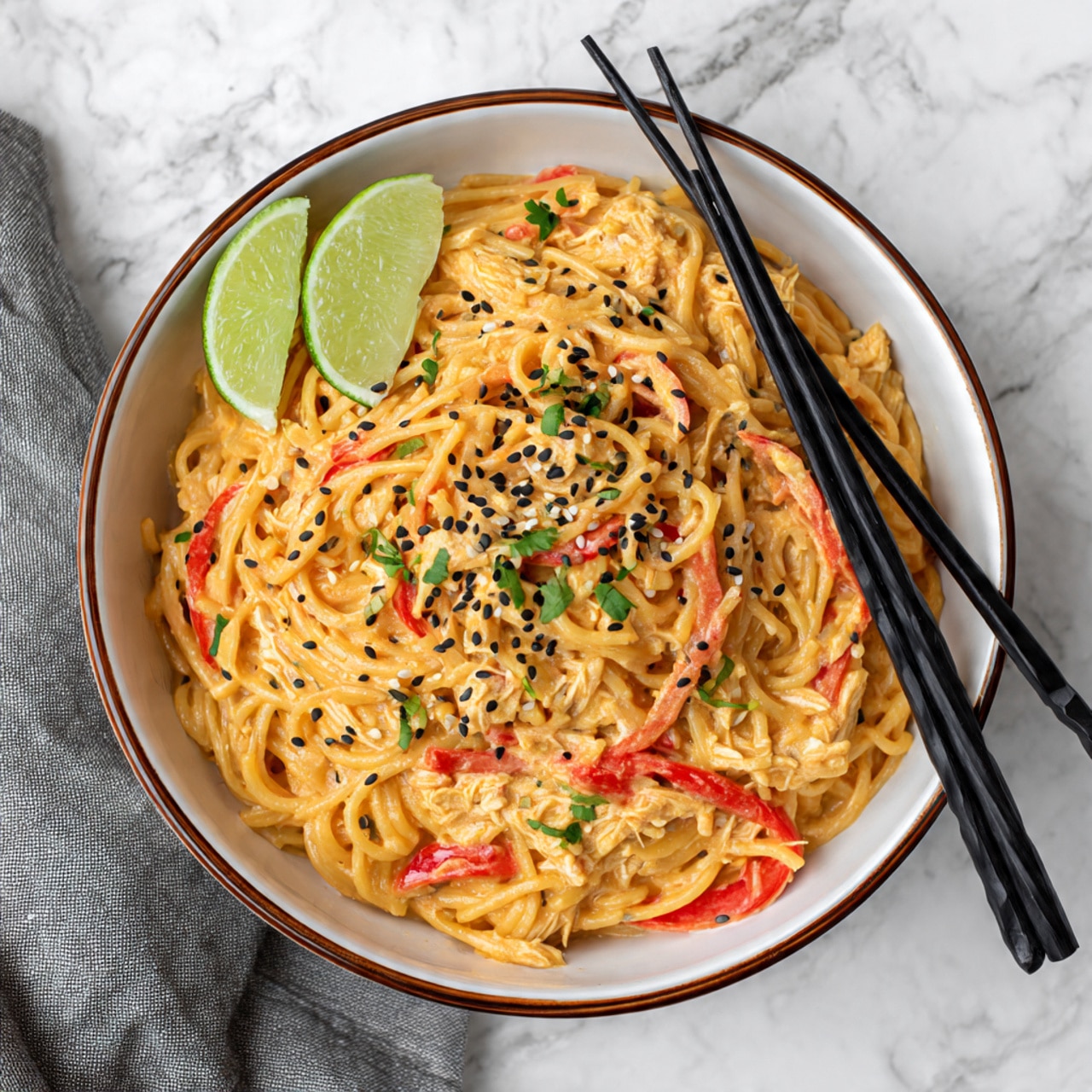 A deep black pan filled with creamy orange sauce coating thin noodles that have a soft, slippery texture, mixed with white chicken strips and thin red bell pepper slices, scattered black sesame seeds adding small dark spots, two bright green lime wedges placed near the top left edge of the pan, the pan set on a light wooden board over a white marbled surface with a gray cloth on the side, a half lime and lime wedge lying on the surface near the pan photo taken with an iphone --ar 4:5 --v 7