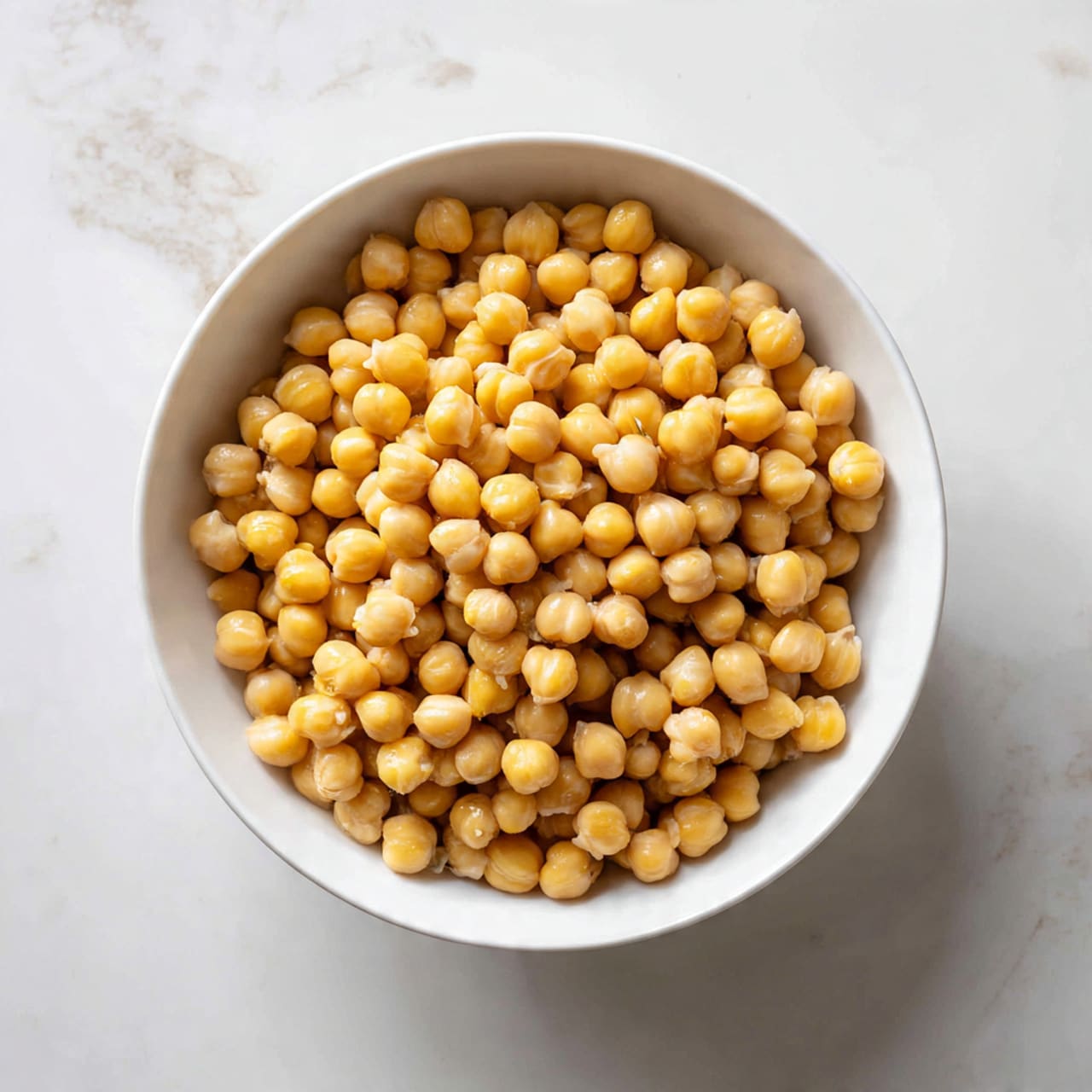 A white bowl filled with a single layer of cooked chickpeas, each chickpea light golden in color, smooth in texture, and round in shape, piled evenly inside the bowl. The bowl sits on a white marbled surface that adds a clean and bright background to the image. The chickpeas fill the bowl nearly to the top, with some slightly overlapping, showing a soft and plump appearance. Photo taken with an iphone --ar 4:5 --v 7
