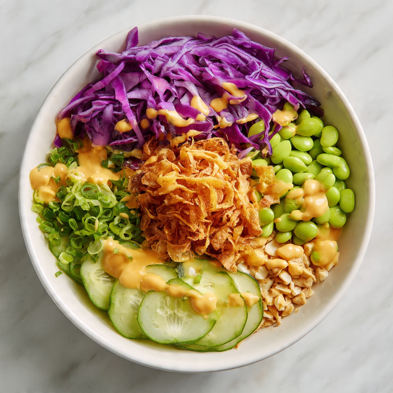 This close-up image shows a vibrant salad with several layers and colors. The bottom layer is fresh, thinly sliced purple cabbage with a rough texture, mixed with bright green edamame beans scattered throughout. On top of this layer, there are thin round slices of light green cucumber visible in different places. Small, crispy golden-brown fried shallots are spread evenly over the top, adding a crunchy texture. There are also pieces of chopped, pale beige cashew nuts sprinkled across the salad. A sprig of fresh green mint leaves adds a fresh touch to one side. The whole salad rests on a white plate, with a white marbled surface visible in the background. The photo taken with an iphone --ar 4:5 --v 7