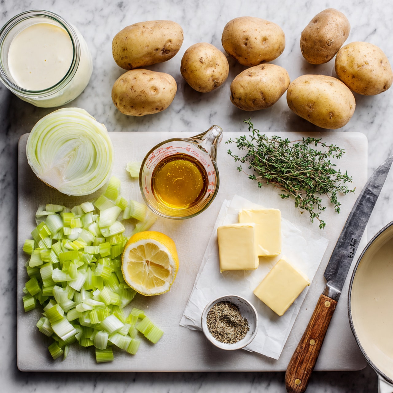 The image shows ingredients for a cooking recipe arranged neatly on a white marbled surface. Four whole potatoes with light brown skin sit near the top center. To their lower right, there are three small rectangular pieces of pale yellow butter on white paper. Below the butter is a glass measuring cup filled with a golden-brown liquid. A small white dish with black pepper and fresh thyme sprigs lies to the right of the measuring cup. On the left side, there is a light green cutting board with chopped pale green celery, sliced white onion, and trimmed pale green-white leeks stacked in layers. A half lemon, cut side up, is placed near the center. A clear glass jar with cream is at the top left. A white pot with a light brown interior and wooden spoon handle is at the bottom right corner. A large silver knife rests on the cutting board. The photo taken with an iphone --ar 4:5 --v 7