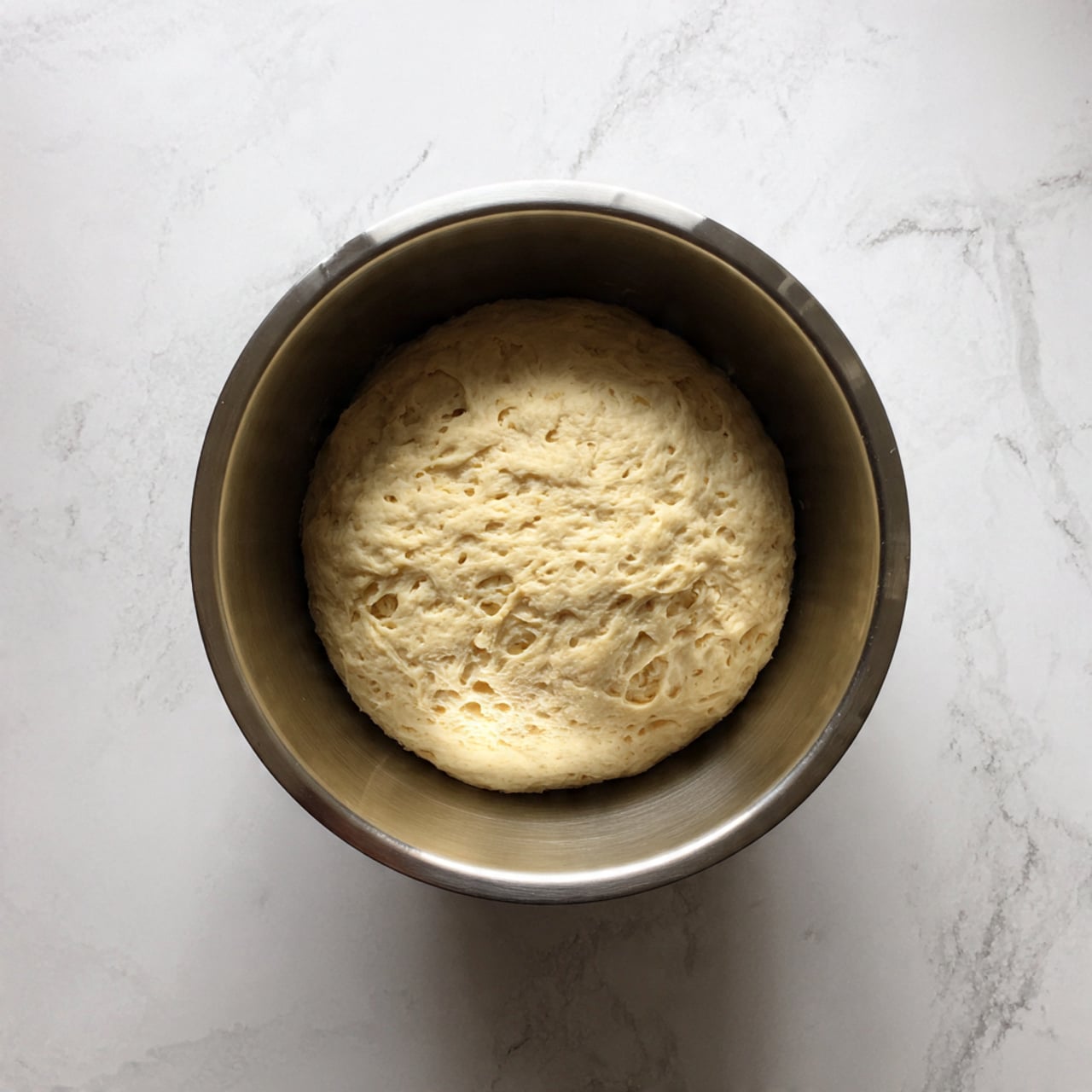 A smooth, round ball of light tan dough sitting inside a metal mixing bowl, showing a soft and slightly bumpy texture with small air pockets, against a white marbled surface background photo taken with an iphone --ar 4:5 --v 7