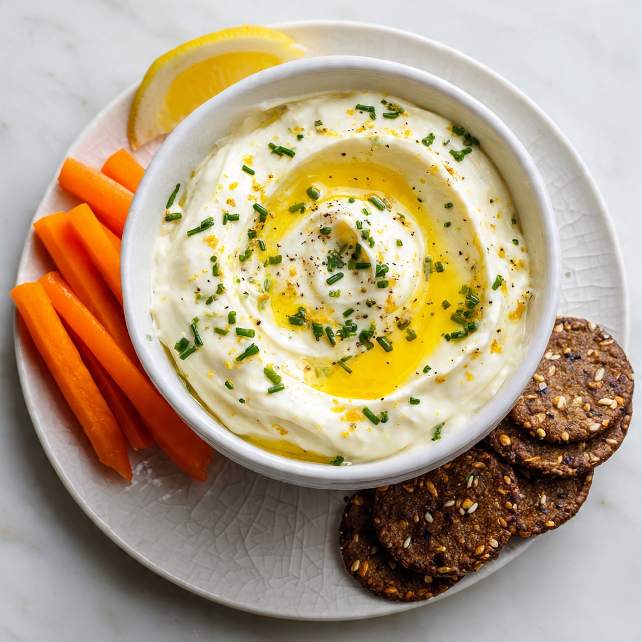 A white plate with brown cracks around the edges holds a thick spread of creamy white dip swirled smoothly in a circular pattern. A golden drizzle of olive oil pools lightly on top of the dip, creating shiny patches. Small pieces of green chopped chives are scattered evenly over the dip. Near the center, a single square cracker with grains and dark spots is partially dipped into the creamy spread. The plate is set on a white marbled surface. photo taken with an iphone --ar 4:5 --v 7