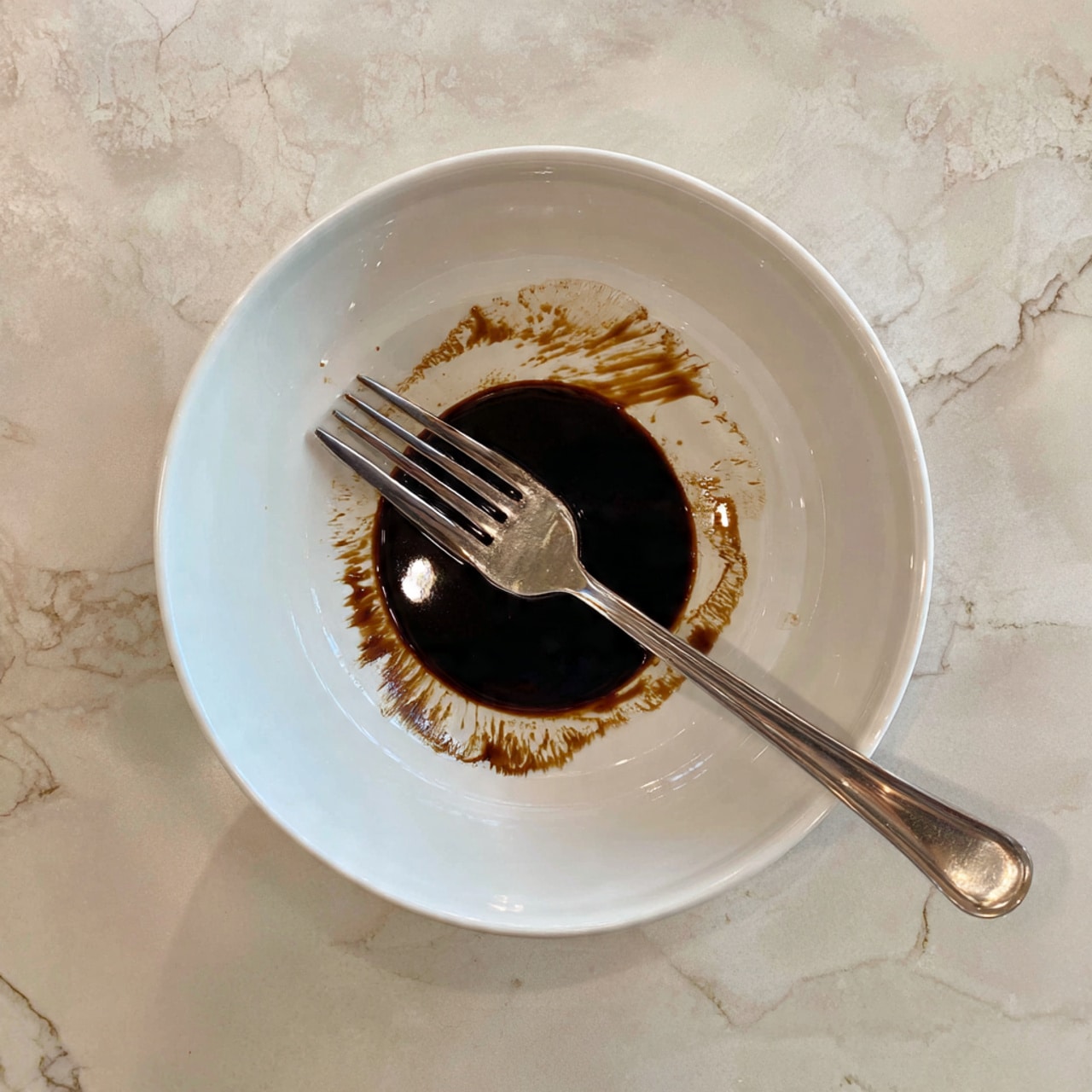 A white bowl sits on a white marbled surface, containing a small amount of dark brown sauce spread unevenly at the bottom. In the center of the bowl lays a silver fork with four tines, partially covered by the sauce. The sauce has a shiny, smooth texture and contrasts against the plain white bowl. Photo taken with an iphone --ar 4:5 --v 7