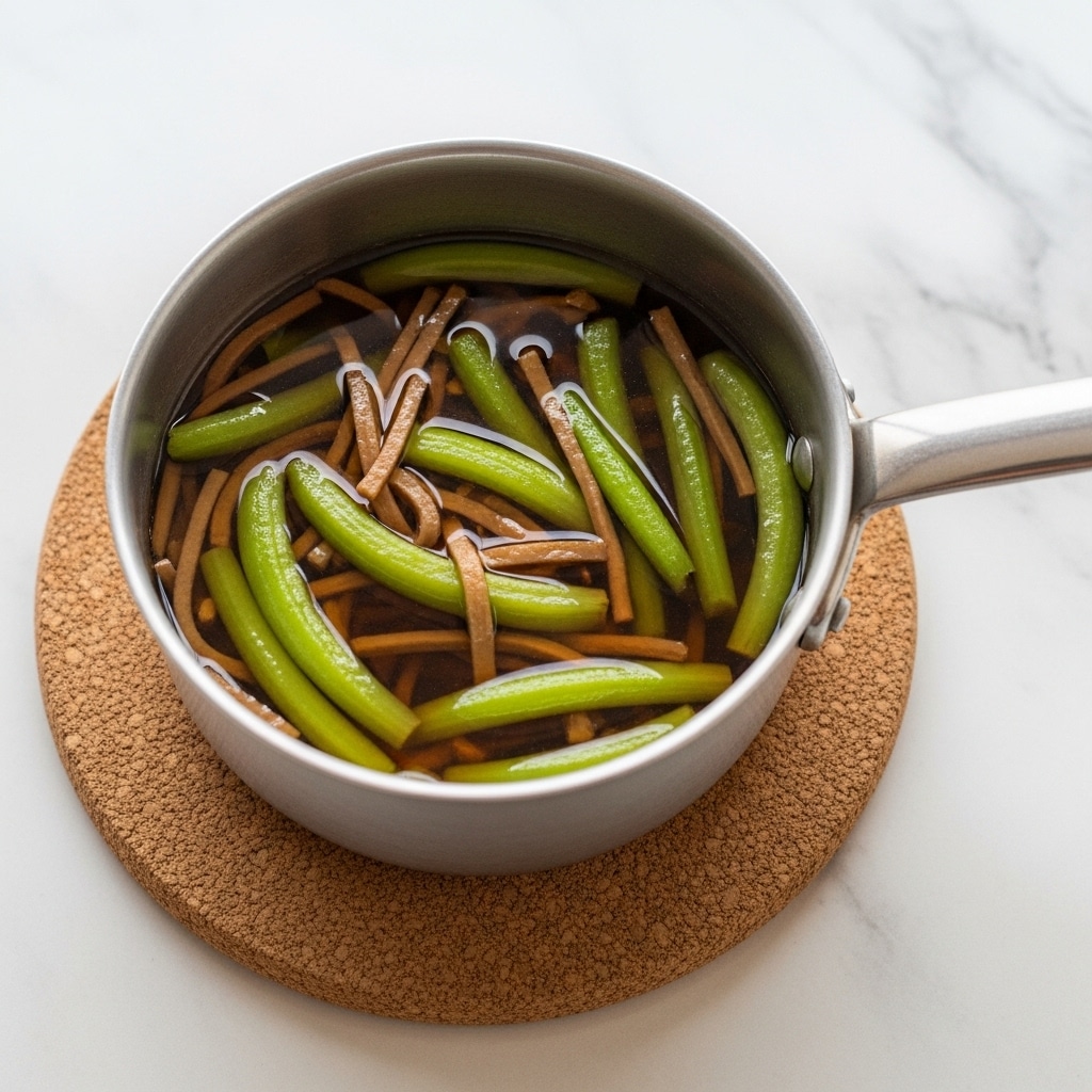 A small silver pot filled with green vegetables and brown thin strips immersed in clear brown broth, placed on a round cork mat against a white marbled surface. The green vegetables are long and slightly curved, mixed with the thin brown strips that add texture in the liquid. The pot handle extends to the right, showing a shiny metallic surface. The broth covers all ingredients, giving a glossy look to the vegetables and strips. The overall color mix is green, brown, and silver with the pot on a soft brown circle mat contrasting the white marbled background. photo taken with an iphone --ar 4:5 --v 7
