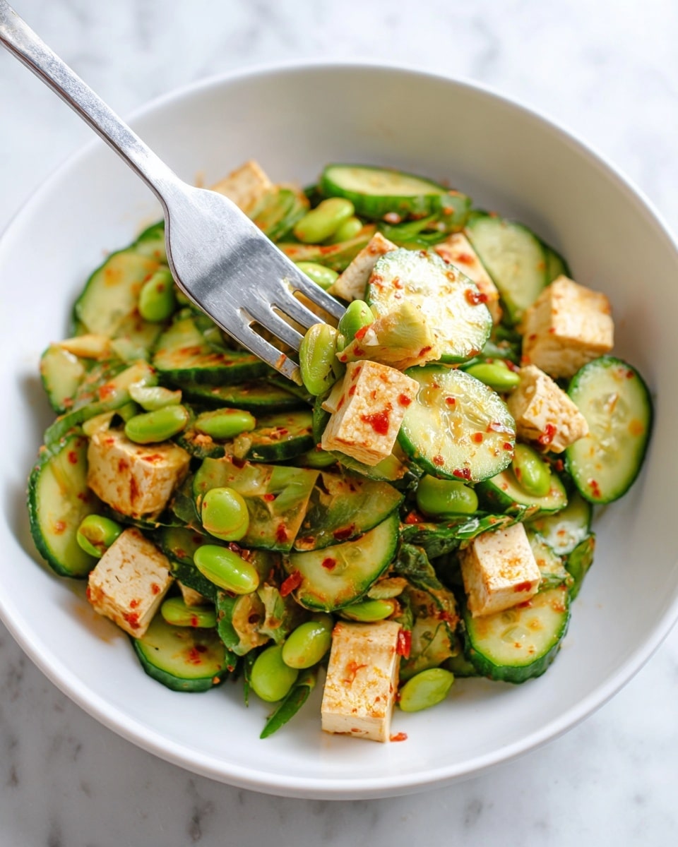 A white bowl filled with a layered salad consisting of thin green cucumber slices scattered throughout, light green edamame beans mixed in, and small cubes of beige tofu with a slightly browned surface. The salad is coated lightly with an orange-red chili sauce that adds speckles of red and a slight glossy texture. A silver fork is holding a mix of the cucumber slices, tofu cubes, and edamame, positioned on the left side near the top of the bowl. The background is a white marbled surface. Photo taken with an iphone --ar 4:5 --v 7
