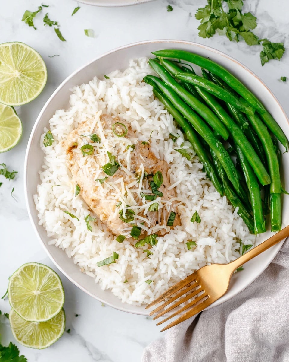 A white plate shows a meal with three main parts: at the bottom, a layer of white rice fills most of the plate with a fluffy, soft texture; on top of the rice in the center, there is a piece of light golden cooked meat garnished with what looks like shredded white cheese and small green chopped herbs. On the right side of the plate, a group of bright green fresh green beans are arranged in a slightly scattered way. A golden fork rests on the right edge of the plate, near the green beans. The background is a white marbled surface with lime slices and a few green herb sprigs nearby. photo taken with an iphone --ar 4:5 --v 7