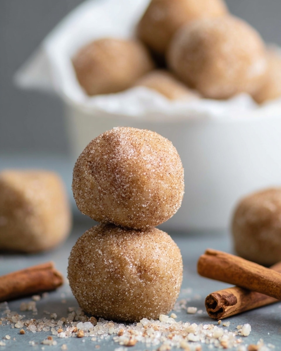 The image shows three round dough balls covered in a cinnamon sugar mix, stacked together on a gray surface with a few cinnamon sticks lying around them. The dough balls have a light brown, slightly rough texture due to the sugar granules and cinnamon coating. In the background, there is a white bowl lined with white paper, filled with more of the same dough balls, slightly blurred to keep focus on the front stack. Some loose sugar and cinnamon granules are scattered around the balls on the surface. photo taken with an iphone --ar 4:5 --v 7