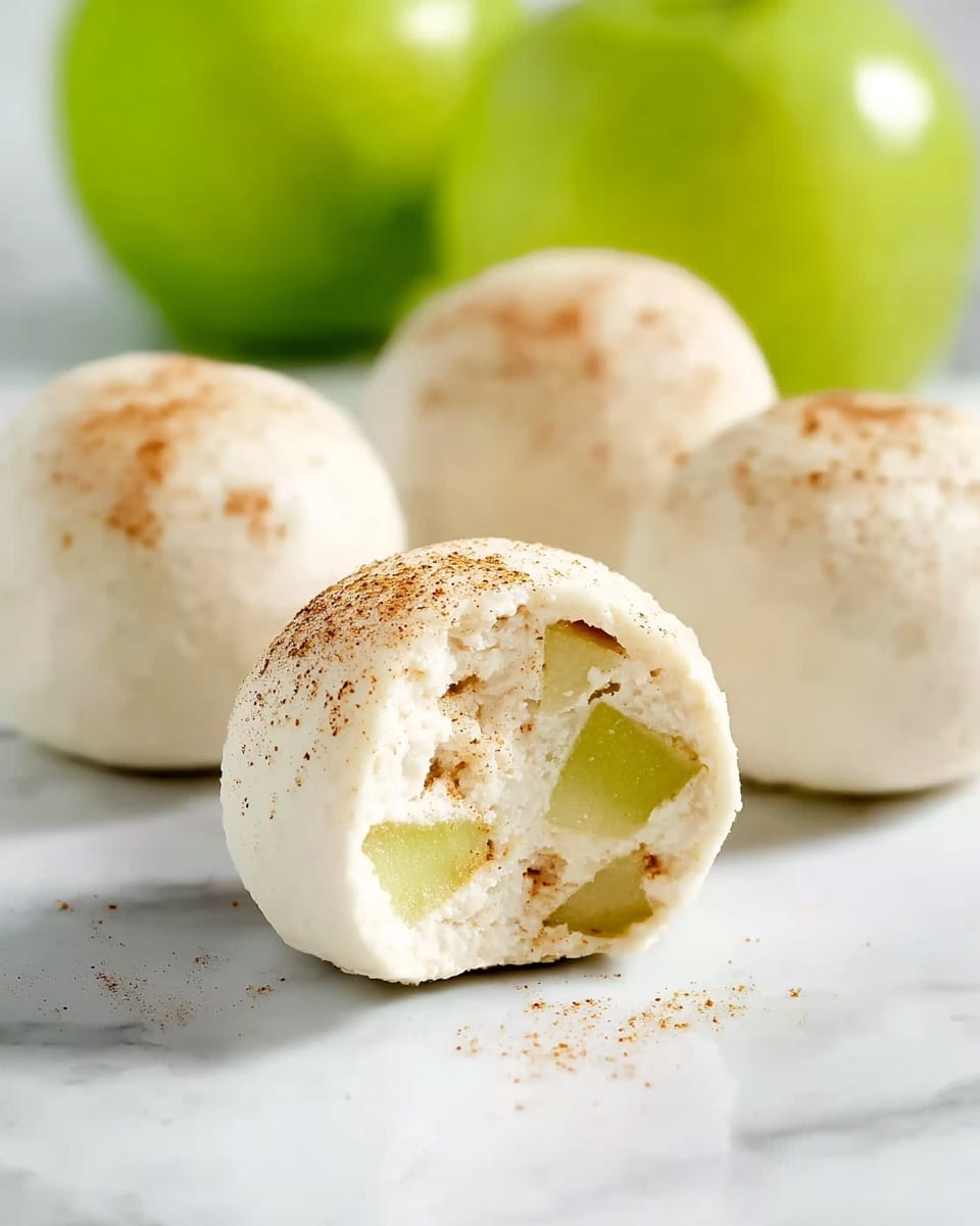 The image shows four round white balls on a white marbled surface, with one ball in the front partially bitten to reveal its soft, creamy inside filled with small pieces of light green apples. The outer layer is smooth, white, and dusted lightly with brown spices, giving a soft matte finish. Behind the balls, there are two bright green apples slightly blurred, adding a fresh hint of color to the background. The photo taken with an iphone --ar 4:5 --v 7