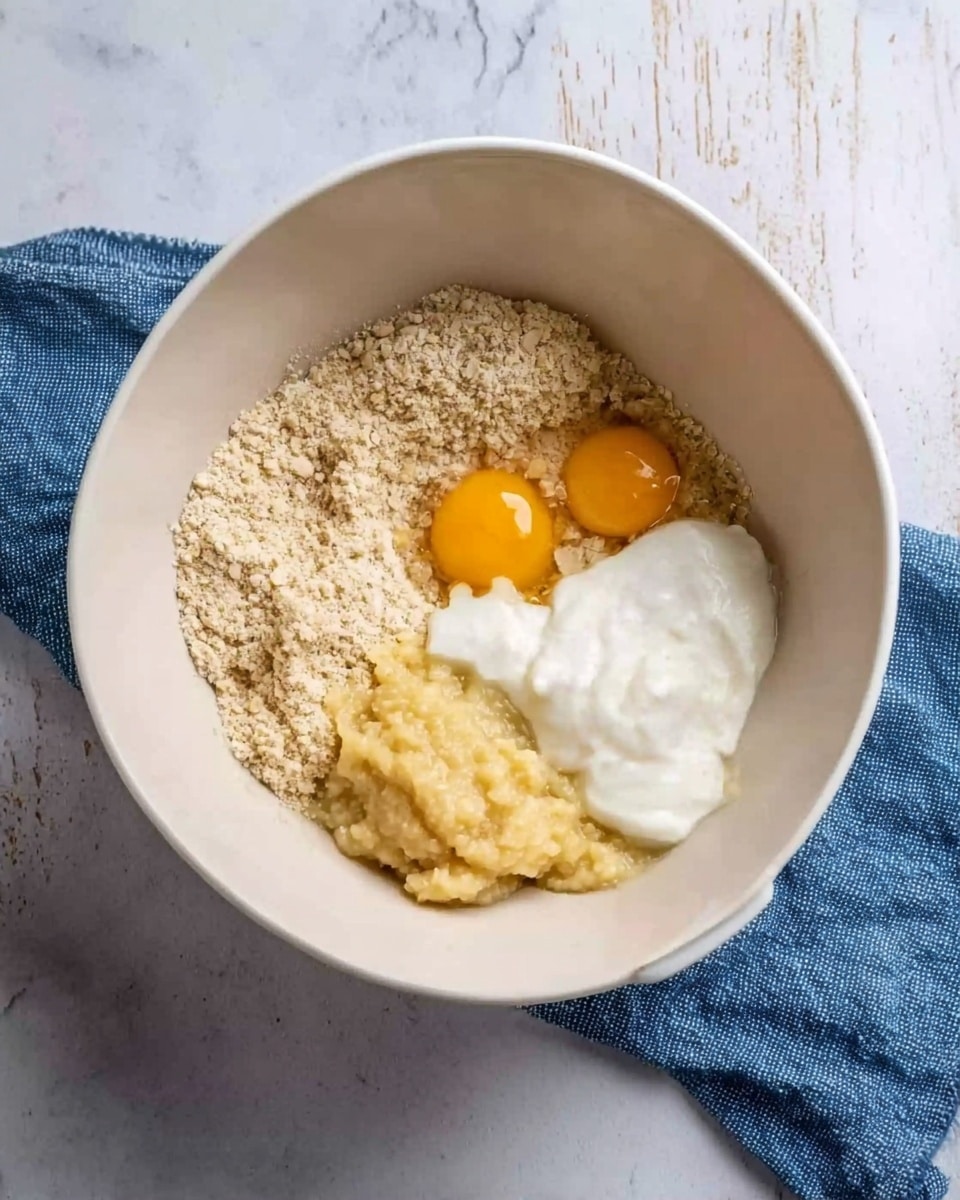 In a white mixing bowl, there are several layers of ingredients placed separately. On the left side, there is a grainy beige mixture, likely flour or oats. On top and slightly to the right of that mixture, two raw egg yolks sit close together with a smooth, thick white cream or yogurt next to them, occupying the upper right section. Below the eggs and cream is a pale yellow mashed mixture, possibly bananas. The bowl is placed on a white marbled surface with a blue cloth nearby. Photo taken with an iphone --ar 4:5 --v 7