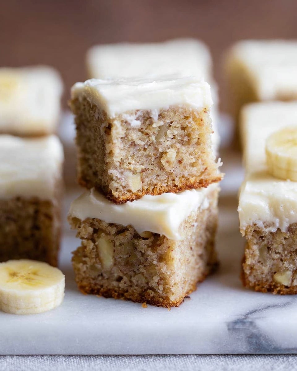 The image shows small square pieces of banana cake stacked on a white marbled surface. Each piece has two layers: a moist, light brown cake base with visible small banana bits and a thick layer of creamy white frosting on top. The pieces are neatly cut and stacked with one piece resting on top of two others, showing the texture inside. A few slices of banana are visible on the white marbled surface near the cake pieces. Photo taken with an iphone --ar 4:5 --v 7