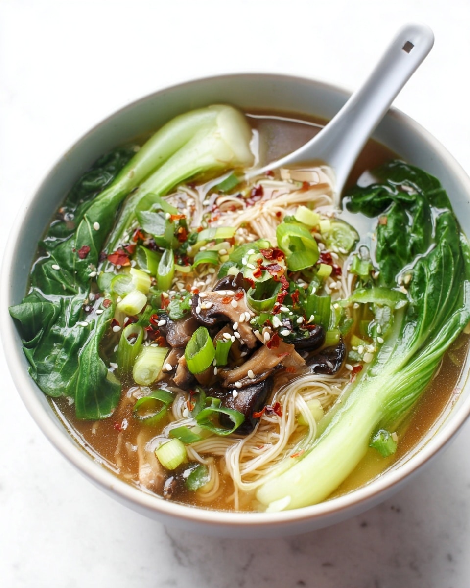 A white bowl filled with clear soup that has thin white noodles layered in the middle, surrounded by bright green bok choy leaves on the sides, dark brown mushroom slices scattered on top, and chopped green onions sprinkled with red chili flakes and white sesame seeds. A white ceramic spoon rests inside the bowl, partially submerged in the broth. The bowl is placed on a white marbled surface. photo taken with an iphone --ar 4:5 --v 7
