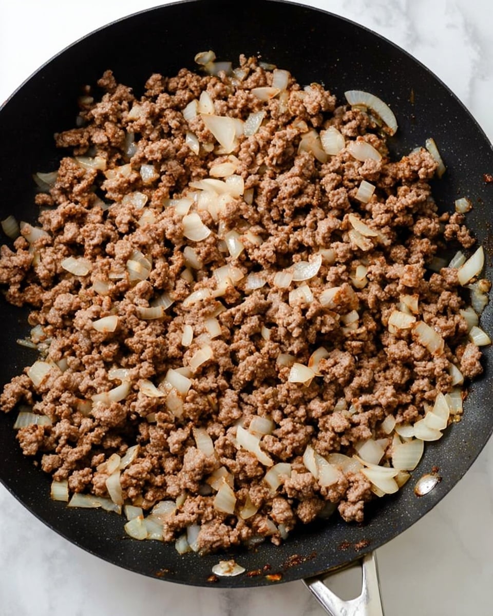 A close-up top view of a black frying pan filled with cooked ground meat mixed with small pieces of translucent white onions. The meat is browned and crumbly with a slightly rough texture, and the onions are evenly spread throughout the meat, showing bits of soft, cooked white. The pan is placed on a white marbled surface with the metal handle partially visible on the right side. photo taken with an iphone --ar 4:5 --v 7