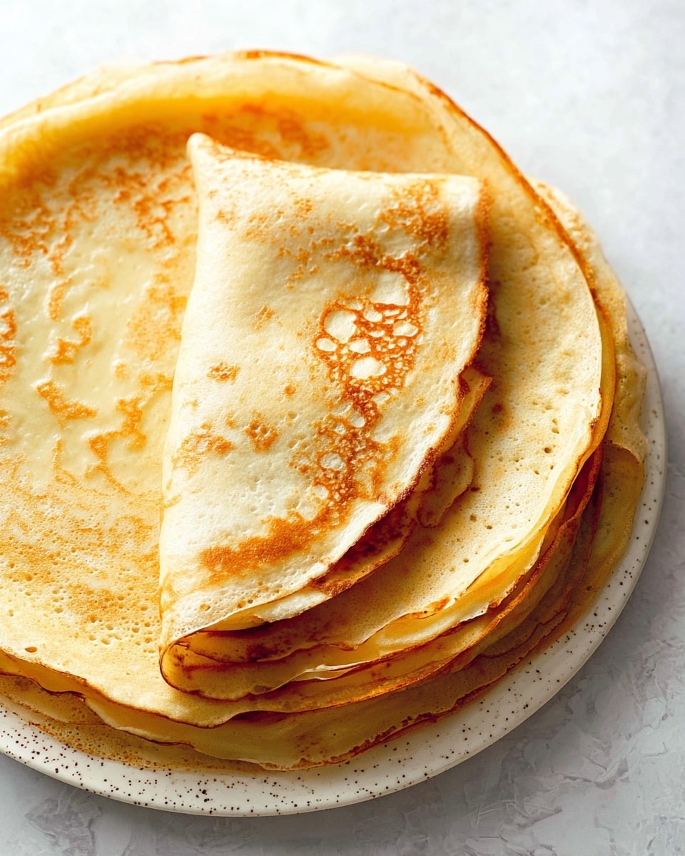 A stack of thin, round crepes with a golden-brown color and light, airy texture is placed on a white plate with small speckles. The top crepe is folded in half, partially covering the crepe underneath, showing soft and slightly crispy edges. The crepes have irregular, delicate patterns of browning, giving them a warm and inviting look. The plate sits on a surface with a white marbled texture. photo taken with an iphone --ar 4:5 --v 7