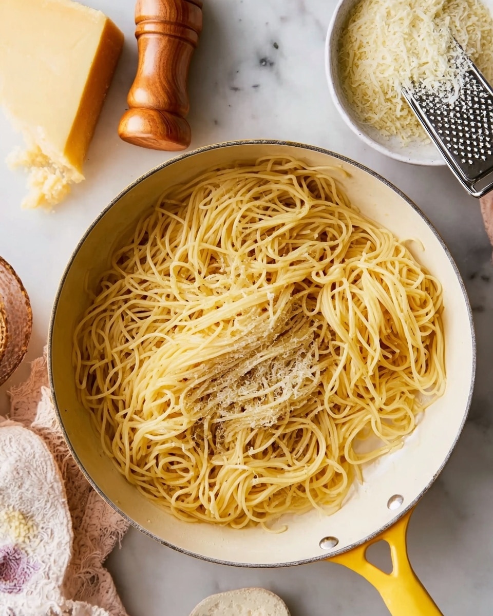 A white pan with a yellow handle filled with cooked spaghetti noodles that look soft and slightly shiny, piled loosely in the center. The pan sits on a white marbled surface. Next to the pan, there is a piece of yellow cheese, a wooden pepper grinder, and a white bowl containing grated cheese with a metal grater resting on top. A woman's hand is holding the pan handle. Photo taken with an iphone --ar 4:5 --v 7