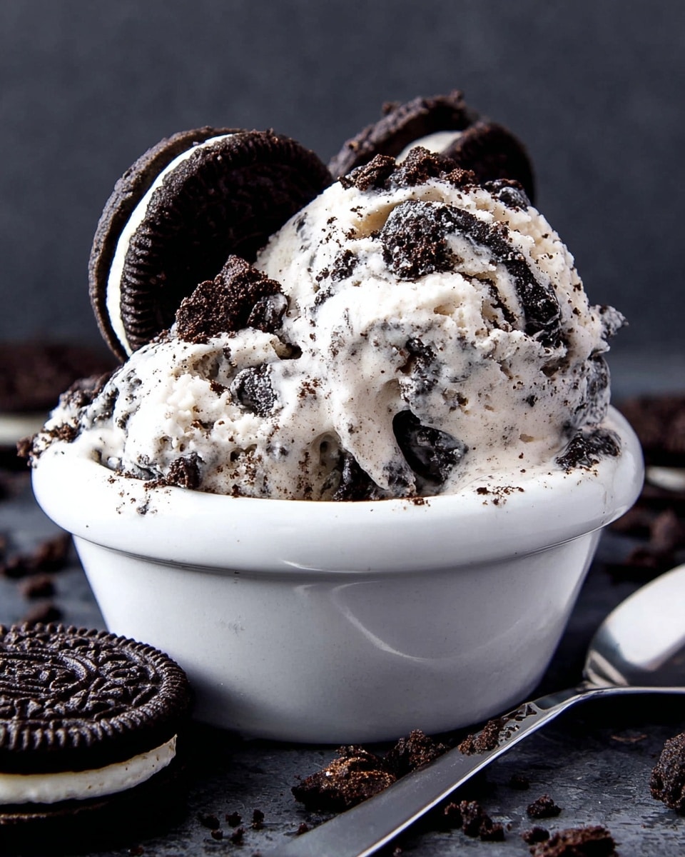 The image shows a small white ceramic bowl filled with two big scoops of cookies and cream ice cream, which is creamy white with many small dark cookie pieces mixed inside. On top, there are extra chunks of crushed dark cookies scattered all around. At the back edge of the bowl, two whole dark cookies with white cream filling are placed upright, partly inside the ice cream. The bowl sits on a dark surface with crumbs of broken cookies scattered around, and a shiny metal spoon lies to the right. The background is a dark gray, and the whole scene is close up, showing thick, rich textures of the ice cream and cookies. Photo taken with an iphone --ar 4:5 --v 7