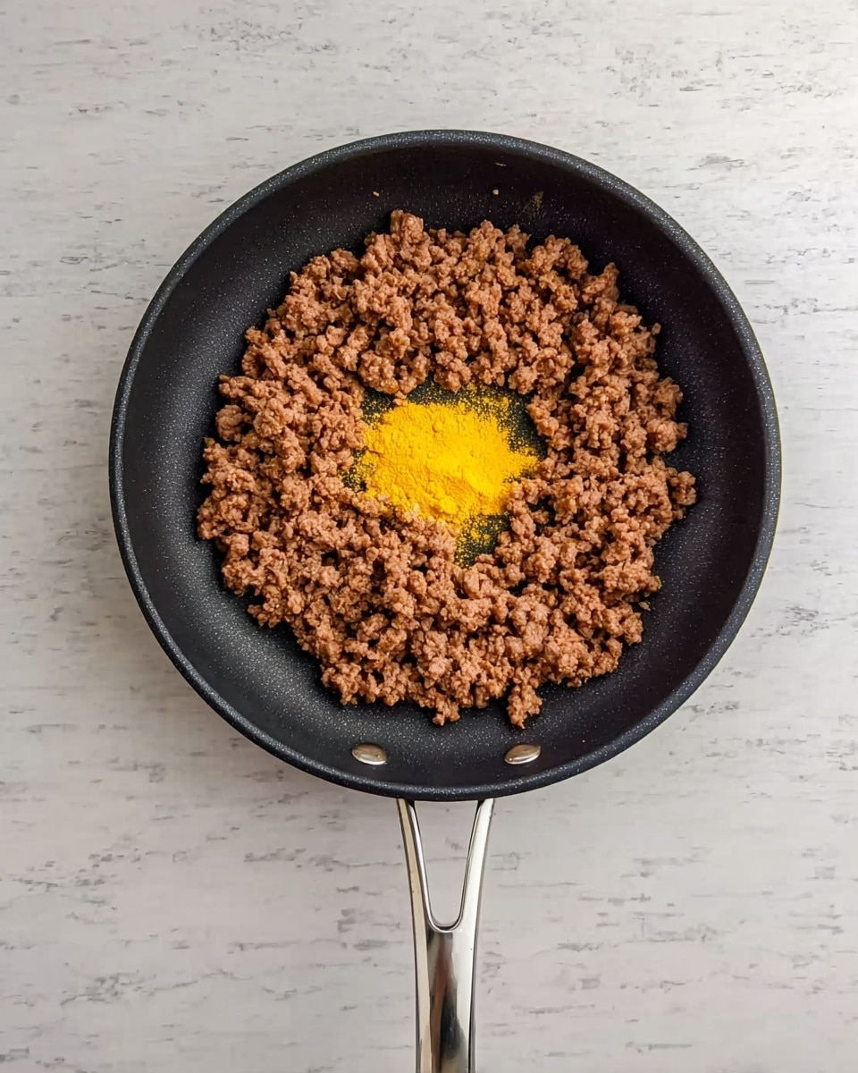 A black frying pan with a silver handle sits on a white marbled surface, filled with a layer of cooked ground meat that is brown and crumbly. In the center of the meat, there is a small pile of bright yellow powder, contrasting strongly with the darker meat around it. The scene is viewed from above, showing the round shape of the pan and the detailed texture of the ingredients inside photo taken with an iphone --ar 4:5 --v 7