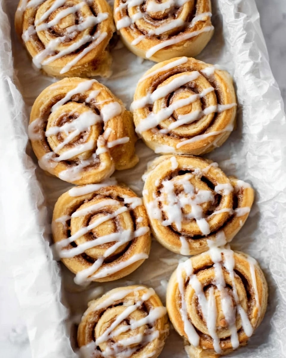 The image shows a group of cinnamon rolls placed in a white tray lined with parchment paper on a white marbled surface. Each roll is round with visible swirls of cinnamon brown filling inside a soft, light golden-brown dough. On top, there are white icing lines drizzled across each cinnamon roll in a slightly messy but even pattern. The rolls have a soft, smooth texture with a slight shine from the icing. photo taken with an iphone --ar 4:5 --v 7