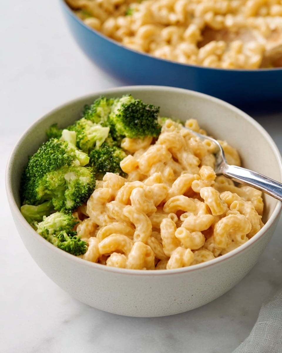 A white bowl sits on a white marbled surface, filled halfway with light golden creamy macaroni pasta showing soft curls and a smooth texture. On one side of the bowl, bright green broccoli florets with a rough, bumpy texture add a fresh contrast. A silver spoon is partially buried in the pasta with its handle resting on the bowl’s edge. In the background, a blue pan containing more creamy macaroni is slightly out of focus. Photo taken with an iphone --ar 4:5 --v 7