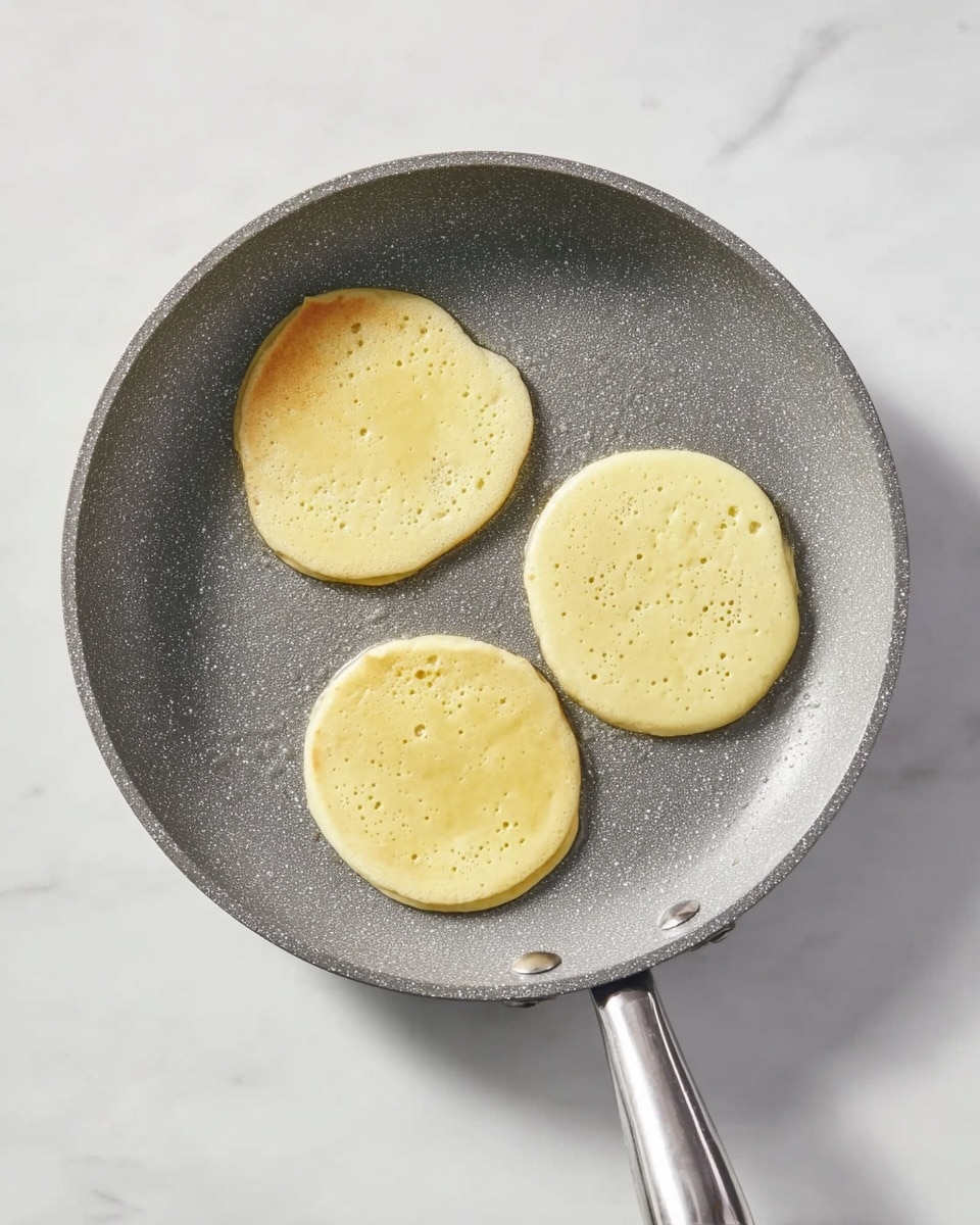 A gray frying pan with a rough texture holds three small pancake-like batter circles cooking evenly on a white marbled surface. Each circle is light yellow with small bubbles appearing on their top sides, showing the batter starting to cook. The pan has a shiny silver handle that extends outward and the white marbled surface underneath adds a clean, bright background. Photo taken with an iphone --ar 4:5 --v 7