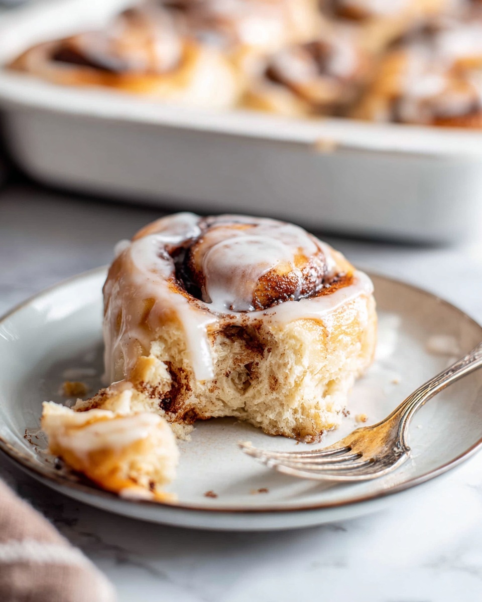 A single cinnamon roll sits on a white plate with soft, fluffy texture and visible layers spiraled with brown cinnamon filling; the top is coated with a shiny white icing that slightly drips down the sides. In front of the roll, there is a torn piece showing the soft inside and cinnamon filling. A fork with traces of icing is placed beside the roll on the right side of the plate. In the background, a white baking tray holds several more cinnamon rolls. The whole scene is set on a white marbled surface. photo taken with an iphone --ar 4:5 --v 7