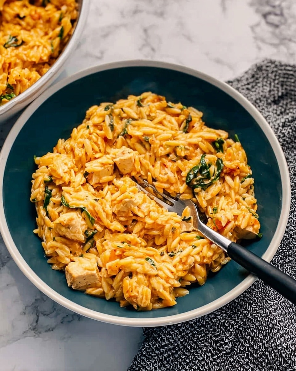 A close-up of a deep white bowl filled with a creamy orzo pasta dish that has a warm orange color mixed with small pieces of white chicken and green spinach. The texture looks rich and slightly saucy, with the pasta and chicken pieces well coated. A fork with a black handle rests inside the bowl on the right side, and the bowl sits on a white marbled surface next to a gray textured cloth. Photo taken with an iphone --ar 4:5 --v 7