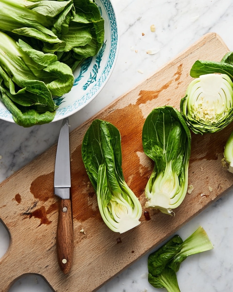 The image shows a wooden cutting board with a light brown color and some water stains, placed on a white marbled surface. On the cutting board, there is a knife with a wooden handle and a silver blade pointing to the right. Two baby bok choy halves with dark green leaves and light green to white stems are positioned near the knife, with one half standing next to the blade and the other slightly tilted. At the far right side of the board, there is a round bok choy base showing its layered pattern. A bunch of fresh whole bok choy with green leaves and pale stems is placed on the marbled surface at the top right corner. On the left side of the image, there is a white bowl with a blue pattern filled with green bok choy leaves, and more green bok choy leaves are scattered partly on the marbled surface near the bowl. photo taken with an iphone --ar 4:5 --v 7