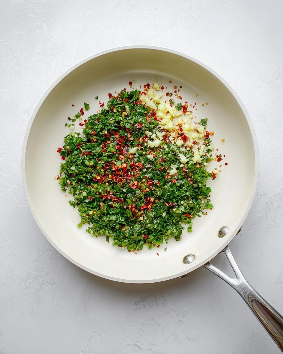 The image shows a white frying pan with a silver handle placed on a white marbled surface. Inside the pan, there is one layer of finely chopped green herbs spread evenly across the bottom. On top of the green herbs, there are small piles of light yellow minced garlic spaced evenly, and scattered red chili flakes with yellow seeds spread mainly around the edges of the herbs but also mixed in. The colors contrast with the white pan, making the ingredients look fresh and vibrant. Photo taken with an iphone --ar 4:5 --v 7