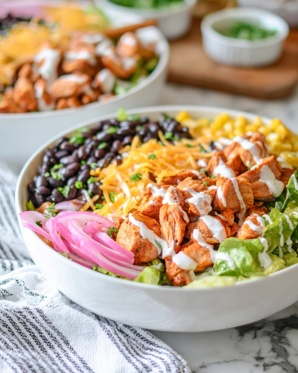 A white bowl filled with a layered salad, starting with a base of green lettuce at the bottom. On top of the lettuce, there are cooked black beans on one side, cooked yellow corn near the back, and reddish-orange cooked chicken pieces on the other side. The chicken is drizzled with a white sauce and sprinkled with shredded orange cheese. There are thin rings of pink pickled onions and chopped green herbs scattered over the top for color and texture. The bowl is set on a white marbled surface with a white and black striped cloth nearby. In the blurry background, there is a second similar bowl. Photo taken with an iphone --ar 4:5 --v 7