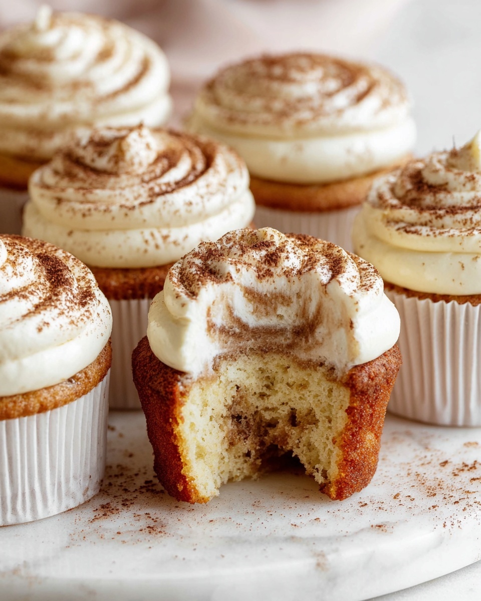A group of seven cupcakes sits on a white marbled surface, with one cupcake at the front showing a bite taken out of it, revealing a light and fluffy inside with a swirl of light brown filling. Each cupcake has two layers: the bottom is a golden brown cake wrapped in a plain white paper liner, and the top is a thick layer of creamy white frosting with a smooth, swirled texture dusted lightly with cocoa powder. The cupcakes are arranged closely together, with soft natural lighting enhancing the texture and color contrast between the frosting and cake. Photo taken with an iphone --ar 4:5 --v 7