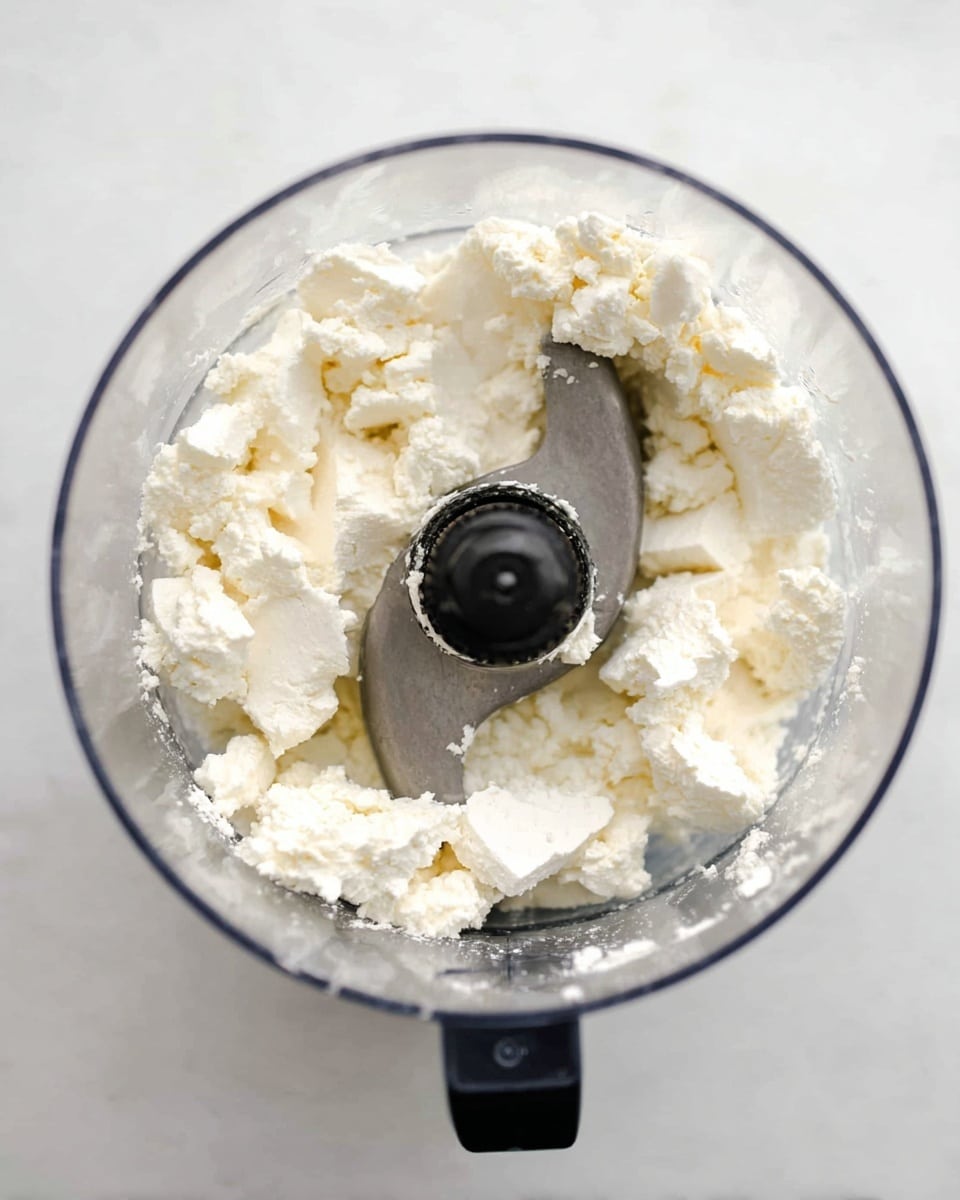 Top view of a clear food processor bowl with uneven white curds or soft cheese pieces inside. The central metal blade is visible with one curved blade coated lightly with some of the white curds. The food processor rests on a white marbled surface, and the whole image has bright and even lighting. photo taken with an iphone --ar 4:5 --v 7