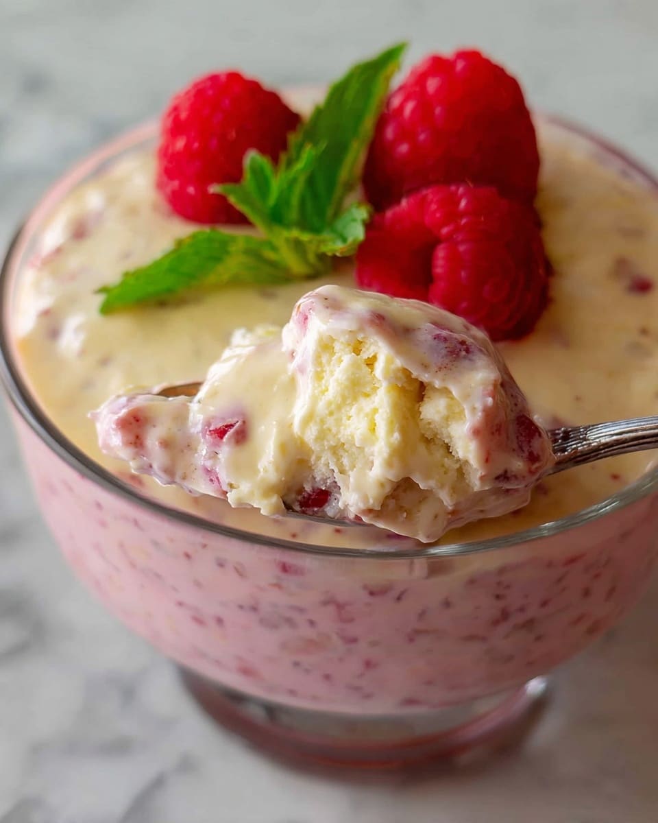 A close-up of a glass bowl filled with a two-layer dessert, the bottom layer is a creamy pink mixture with visible small seeds and fruit bits, topped with a smooth, pale yellow creamy layer. On top, there are three fresh red raspberries and a green mint leaf for garnish. A spoon holds up some of the pink layer mixed with a chunk of pale yellow cream, showing a soft and creamy texture. The bowl sits on a white marbled surface. Photo taken with an iphone --ar 4:5 --v 7