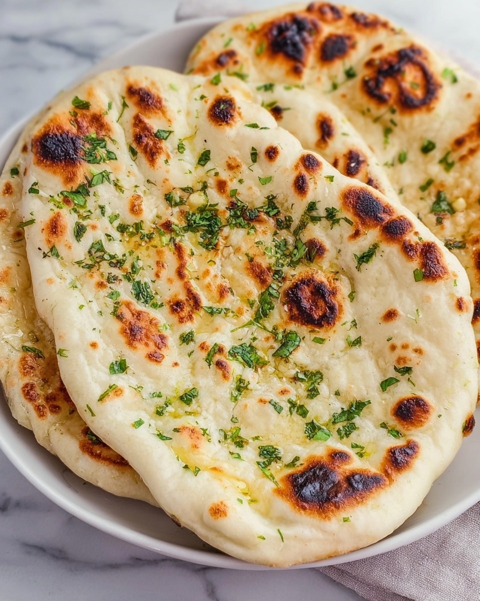 Two pieces of naan bread rest on a white plate, each piece showing a soft, slightly puffed texture with visible air bubbles and golden brown charred spots scattered across their surface. The tops are sprinkled with chopped green herbs and small bits of garlic, adding a fresh look. The edges are thicker and a creamy off-white color with some darker toasted areas. The white marbled surface under the plate brightens the scene. photo taken with an iphone --ar 4:5 --v 7