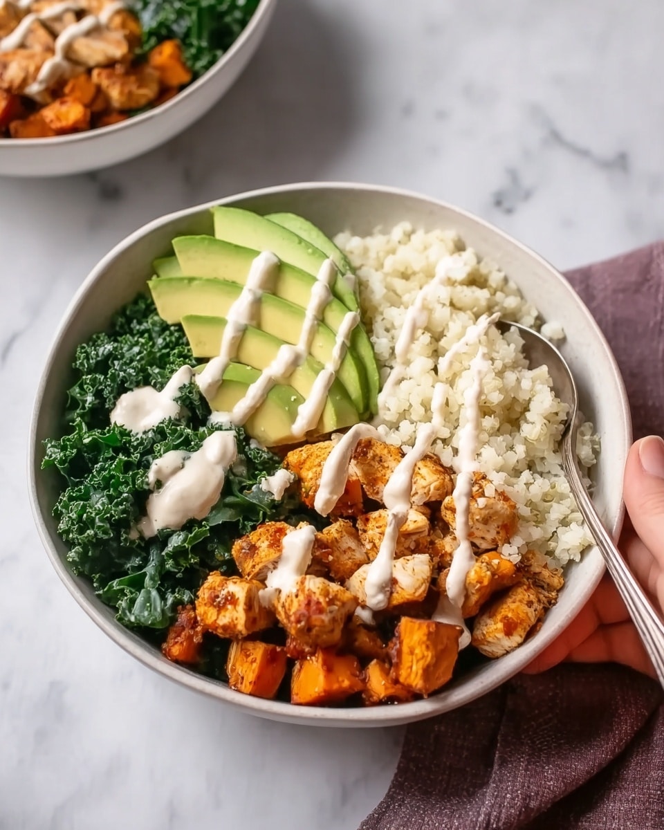 The image shows a white bowl filled with a healthy meal placed on a white marbled surface. Inside the bowl, there are four distinct layers: on the left side, a dark green, leafy kale layer with a slightly rough texture; next to it, thin slices of light green avocado curved neatly; then a fluffy, white cauliflower rice layer with a grainy texture sits on the right. The bottom layer along the front half of the bowl contains small, orange-brown roasted sweet potato cubes and light brown cooked chicken pieces. All layers are topped with thin, creamy white sauce drizzled in lines across the bowl. A shiny silver spoon rests inside the bowl on the right side, and a woman’s hand is holding the bowl slightly from the top left. Another similar bowl is partially visible blurred in the background. Photo taken with an iphone --ar 4:5 --v 7