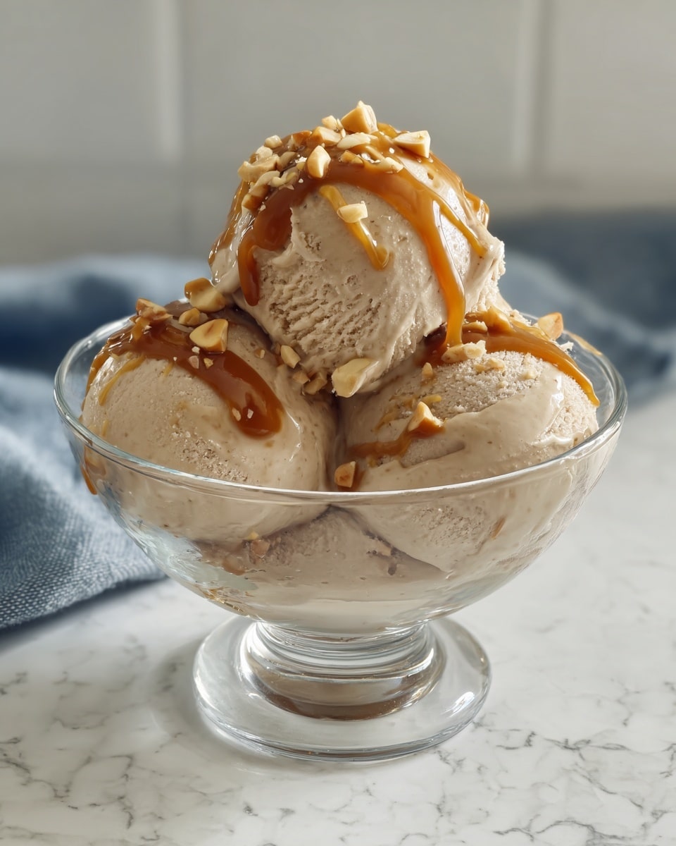 A clear glass bowl holds three scoops of light brown ice cream with a smooth but slightly textured surface. The top scoop is drizzled generously with thick caramel sauce that runs down the sides and is sprinkled with chopped light tan nuts. The bowl sits on a white marbled surface with a soft blurred background. Photo taken with an iphone --ar 4:5 --v 7